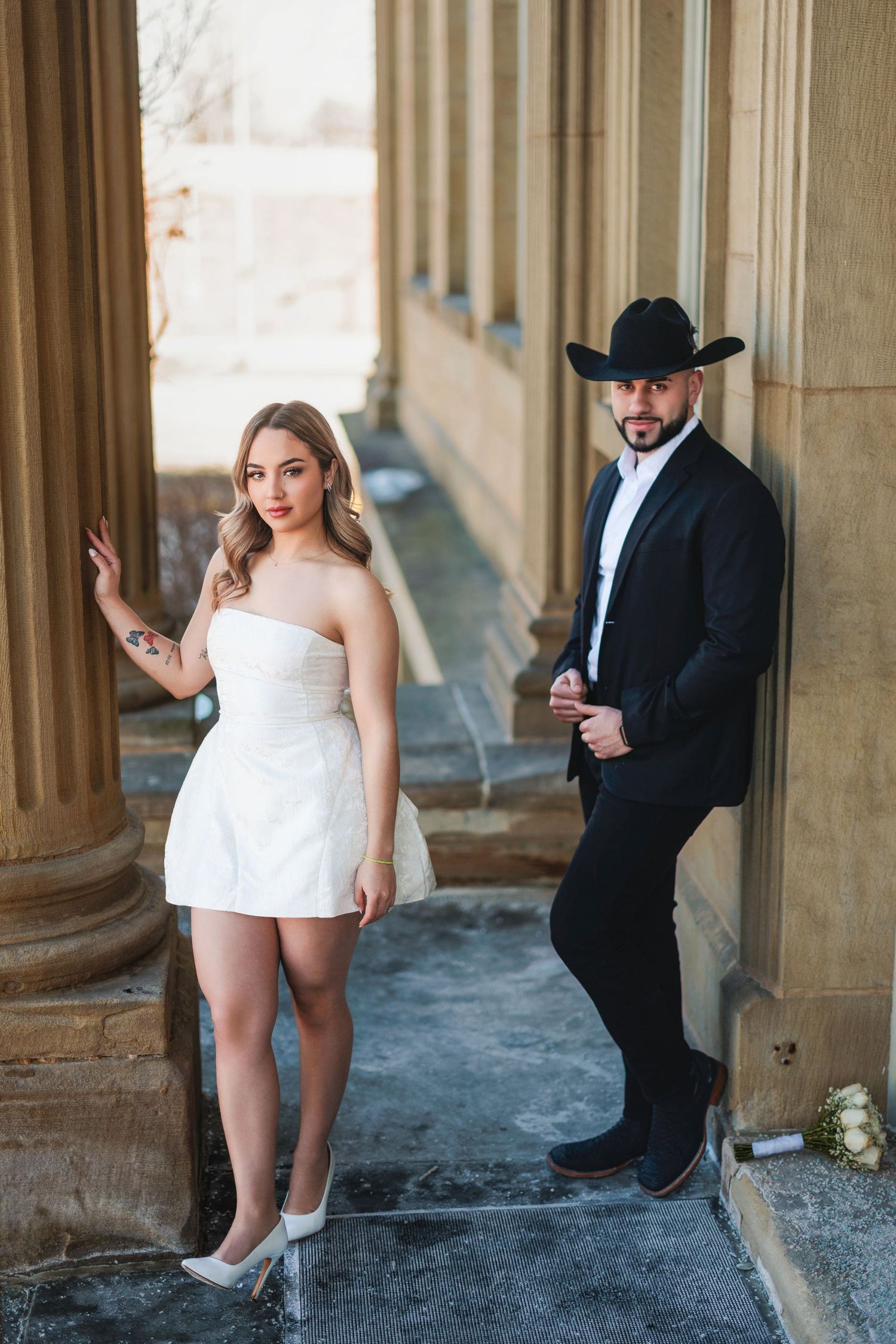 Couple in contrasting formal attire pose between elegant stone columns, her in white dress and him in black suit with cowboy hat.