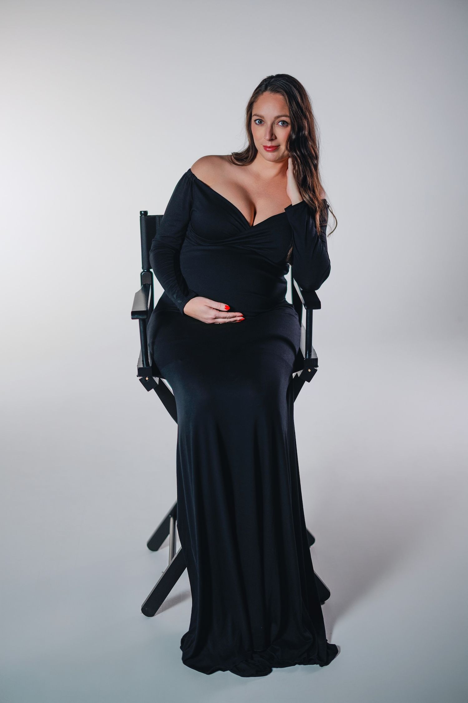 A figure poses in an elegant black off-shoulder maternity gown while seated in a dark chair against a light gray backdrop.