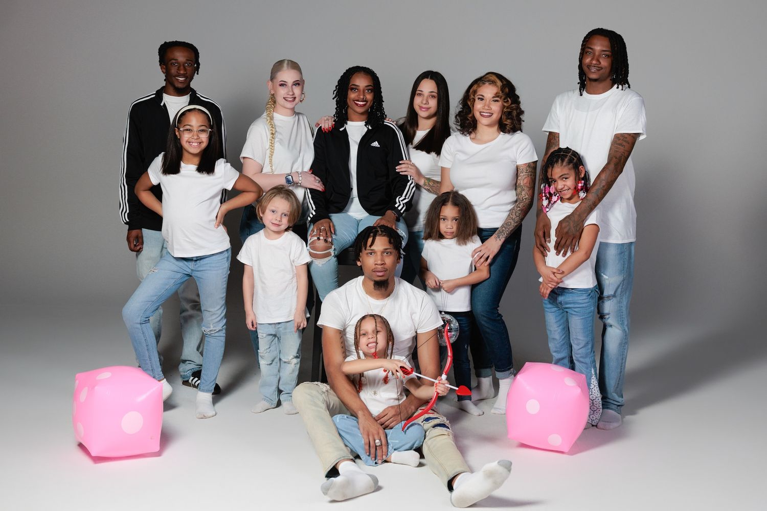 A group poses together in a studio setting wearing casual white and denim outfits with pink accent props in the background.