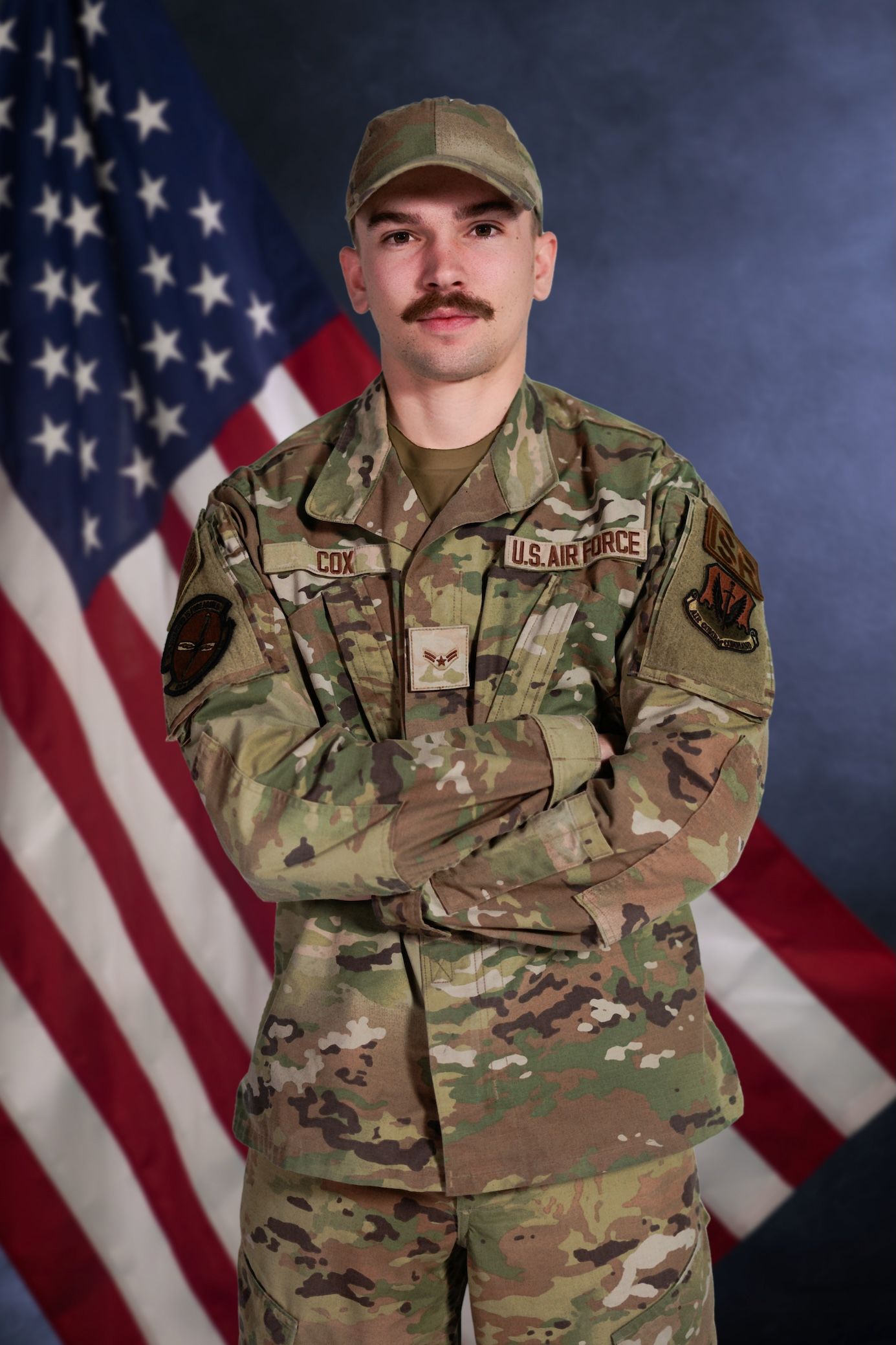 A service member in camouflage uniform stands at attention in front of an American flag.