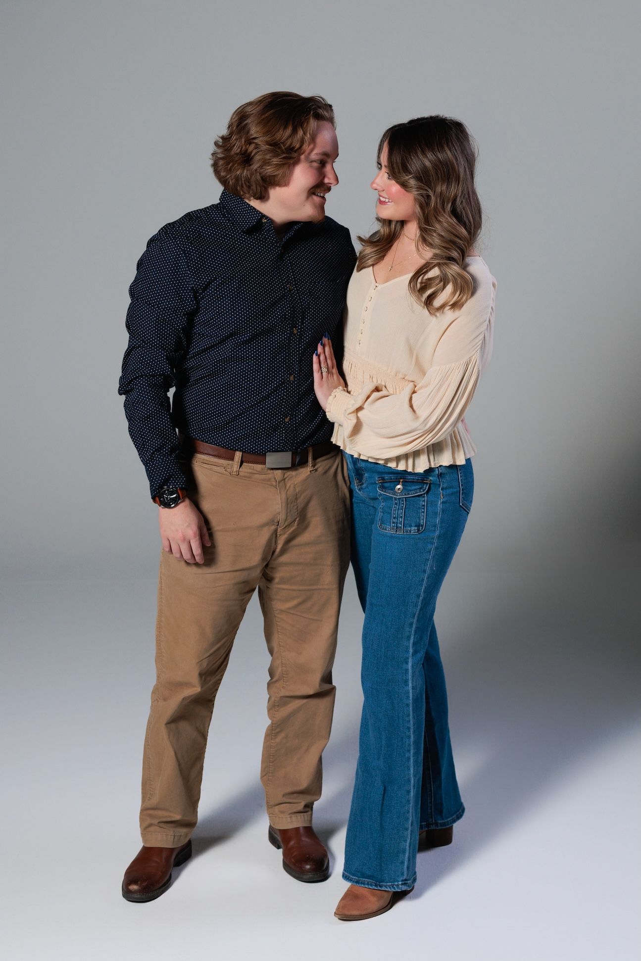 A couple in casual sweaters and jeans embracing and smiling at each other against a gray studio backdrop.