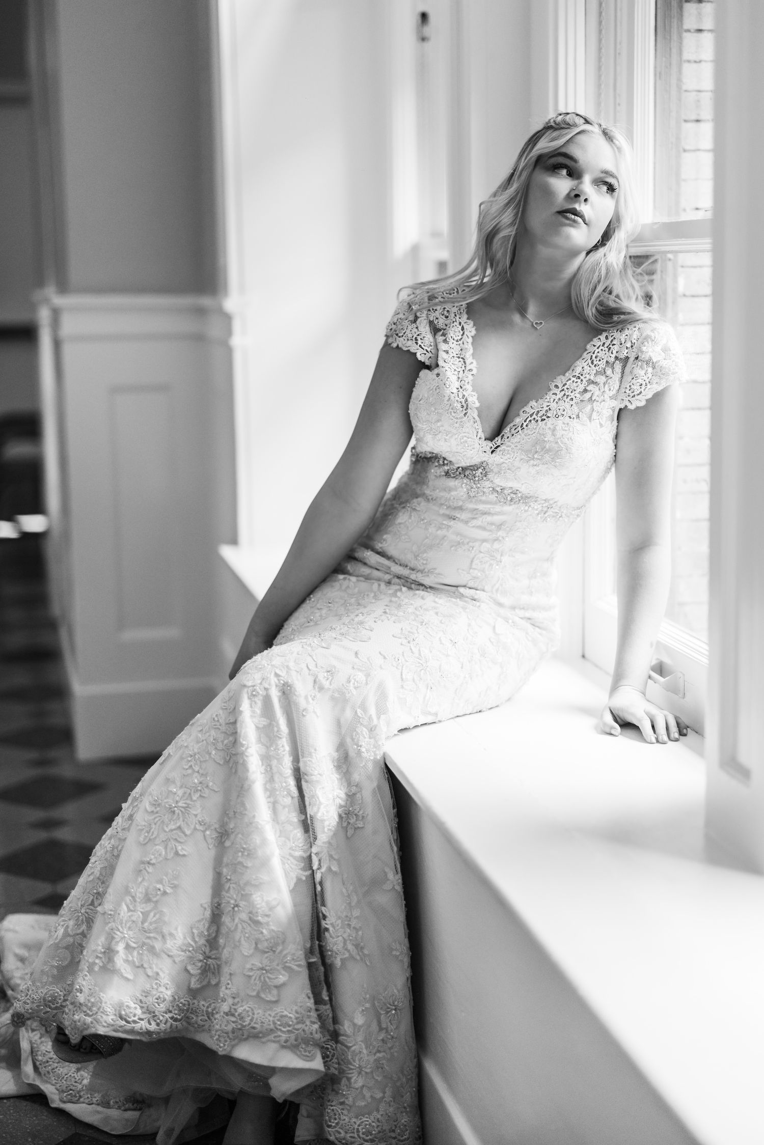 A black and white photograph shows a bride in an elegant lace wedding dress with cap sleeves seated by a bright window.