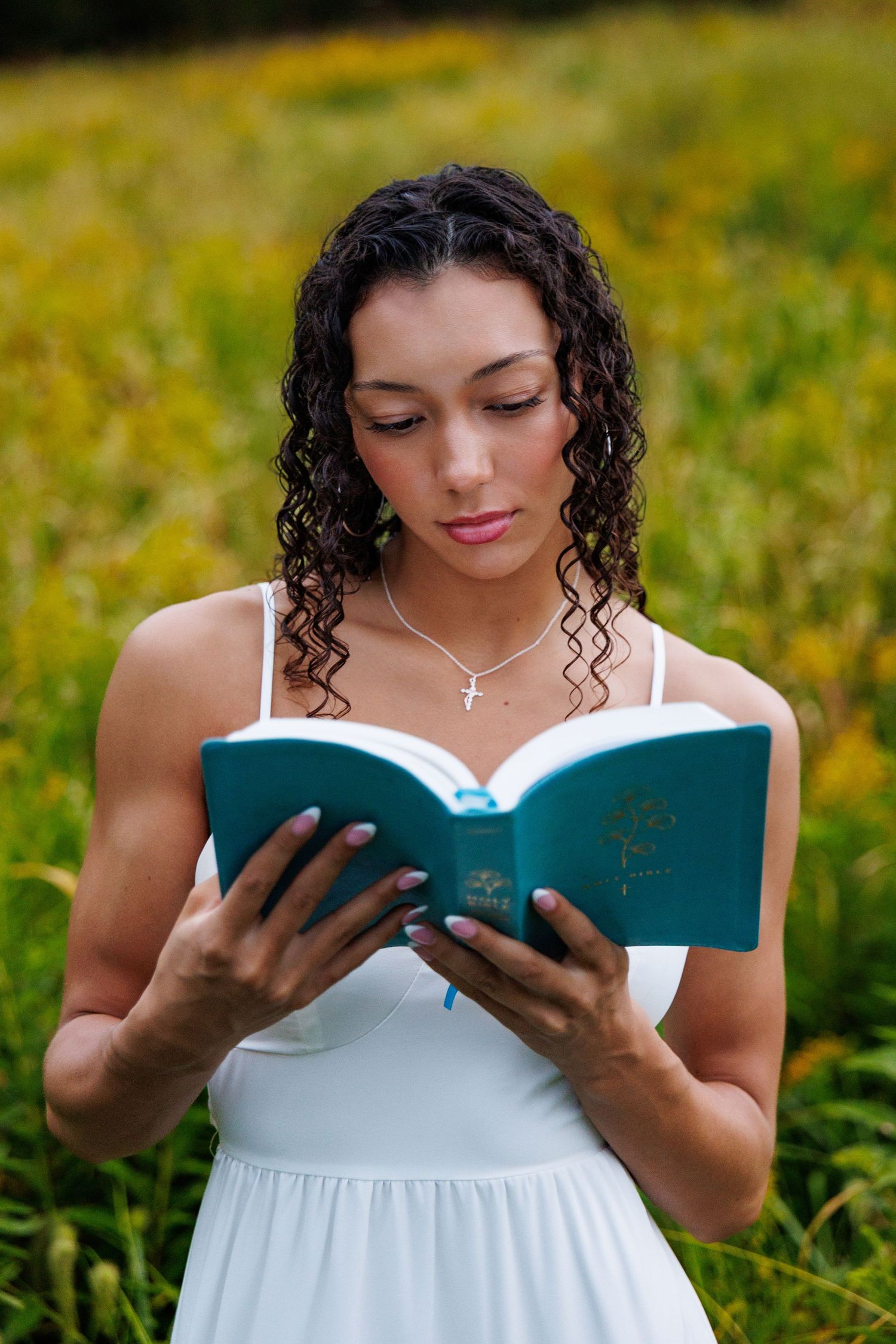 A person in a white dress reads from a book while standing in a lush green field on a sunny day.