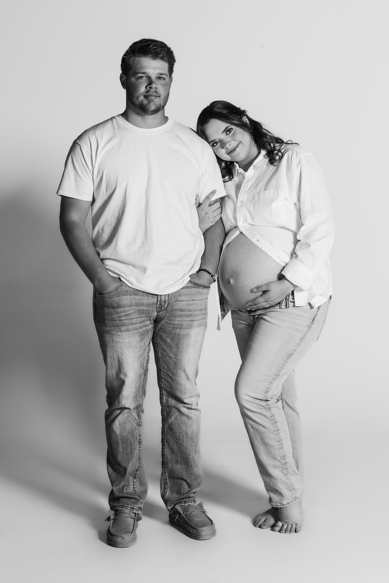 Black and white maternity photo of couple in casual clothes standing together against light backdrop.