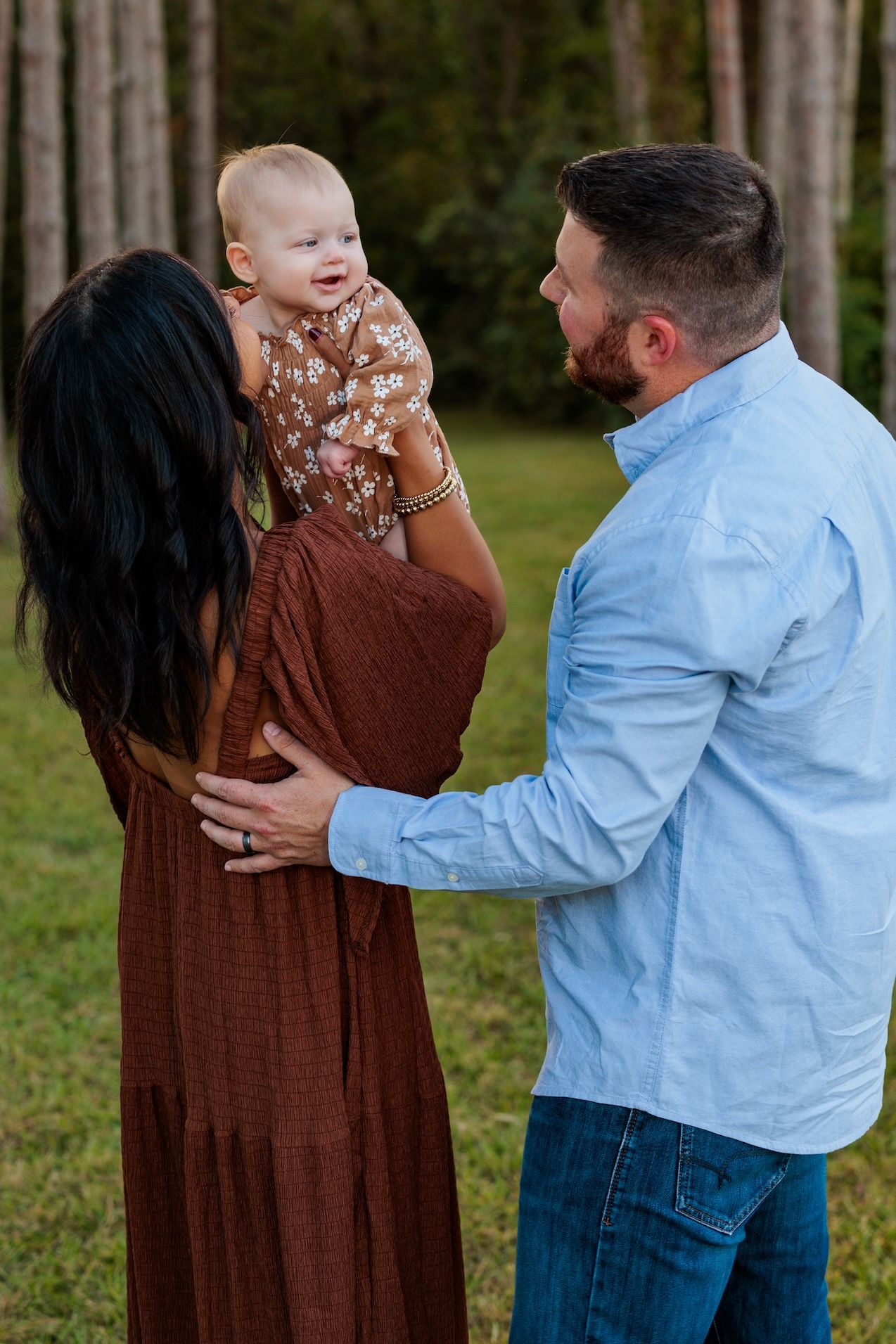 A family shares a joyful moment together outdoors in a wooded setting, with the baby wearing a floral outfit.