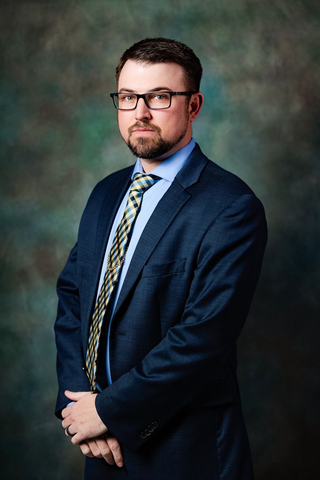 Professional portrait of a businessman in a navy suit, patterned tie and glasses against a mottled dark green backdrop.