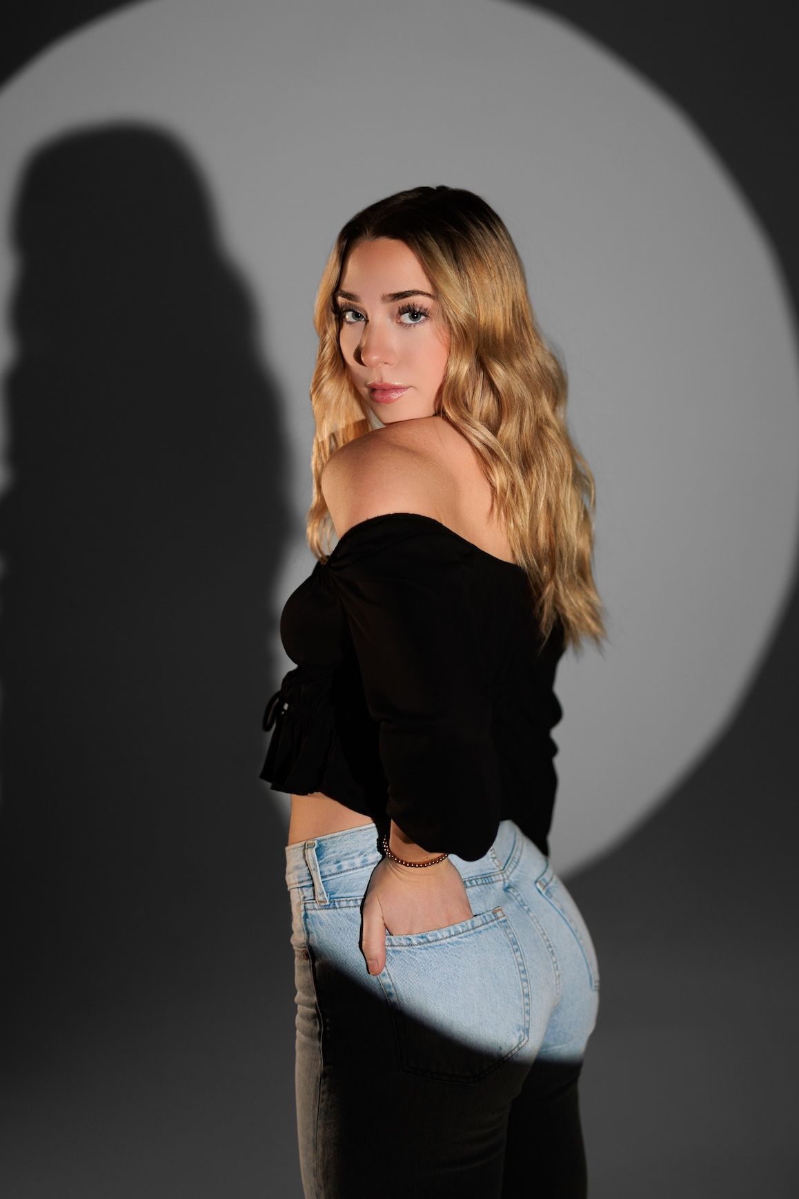 Dramatic studio portrait with deep shadows featuring black off-shoulder top and light denim jeans against circular spotlight.