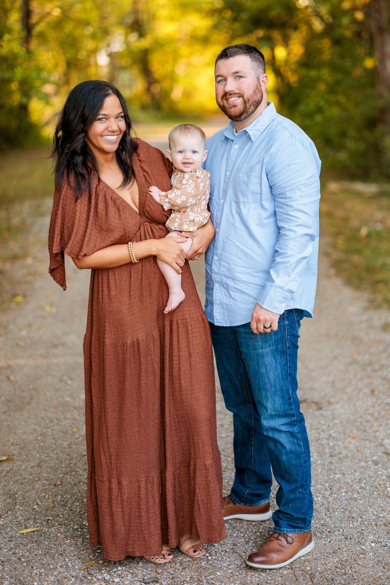 A family poses together outdoors on a gravel path surrounded by golden autumn foliage in the background.