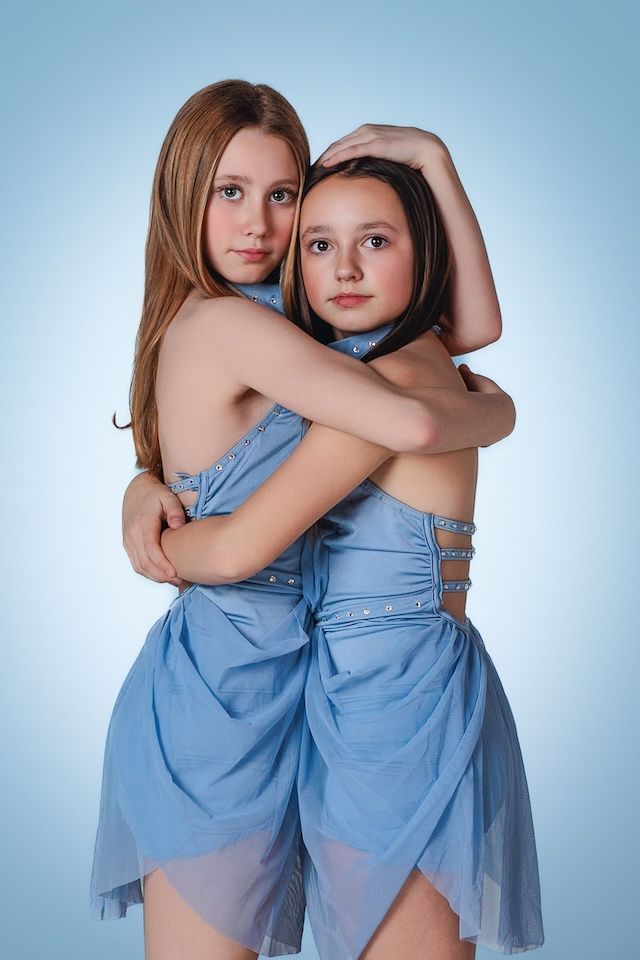 Two dancers in flowing light blue dresses pose together against a blue background.