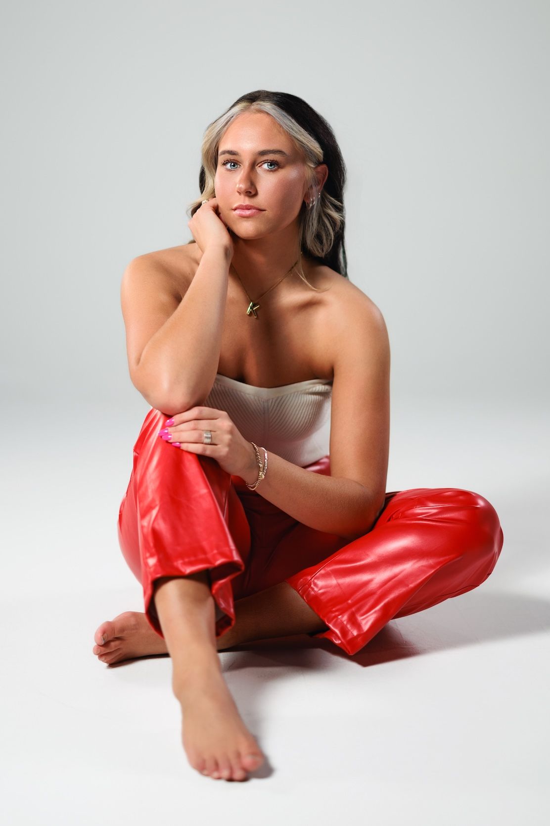 Stylish portrait against white background wearing red leather pants and white top in contemplative seated pose.
