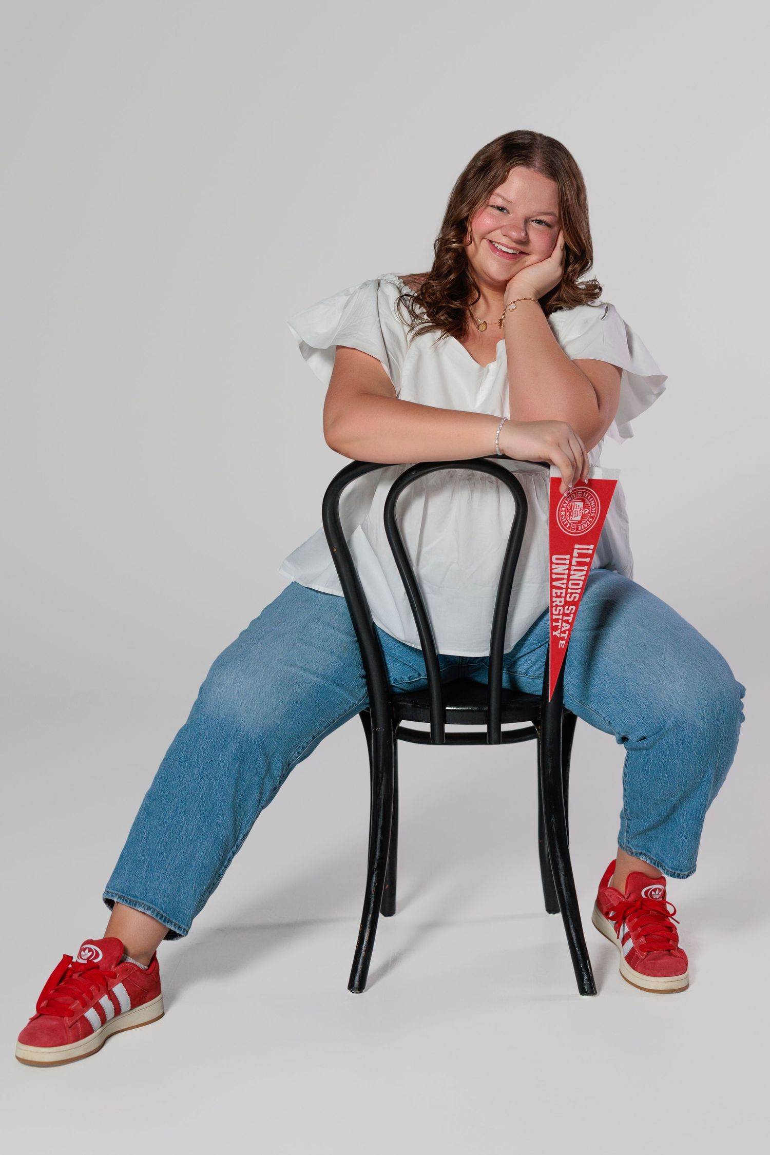 Person in white top and jeans sits casually on black bentwood chair against white background wearing red sneakers.