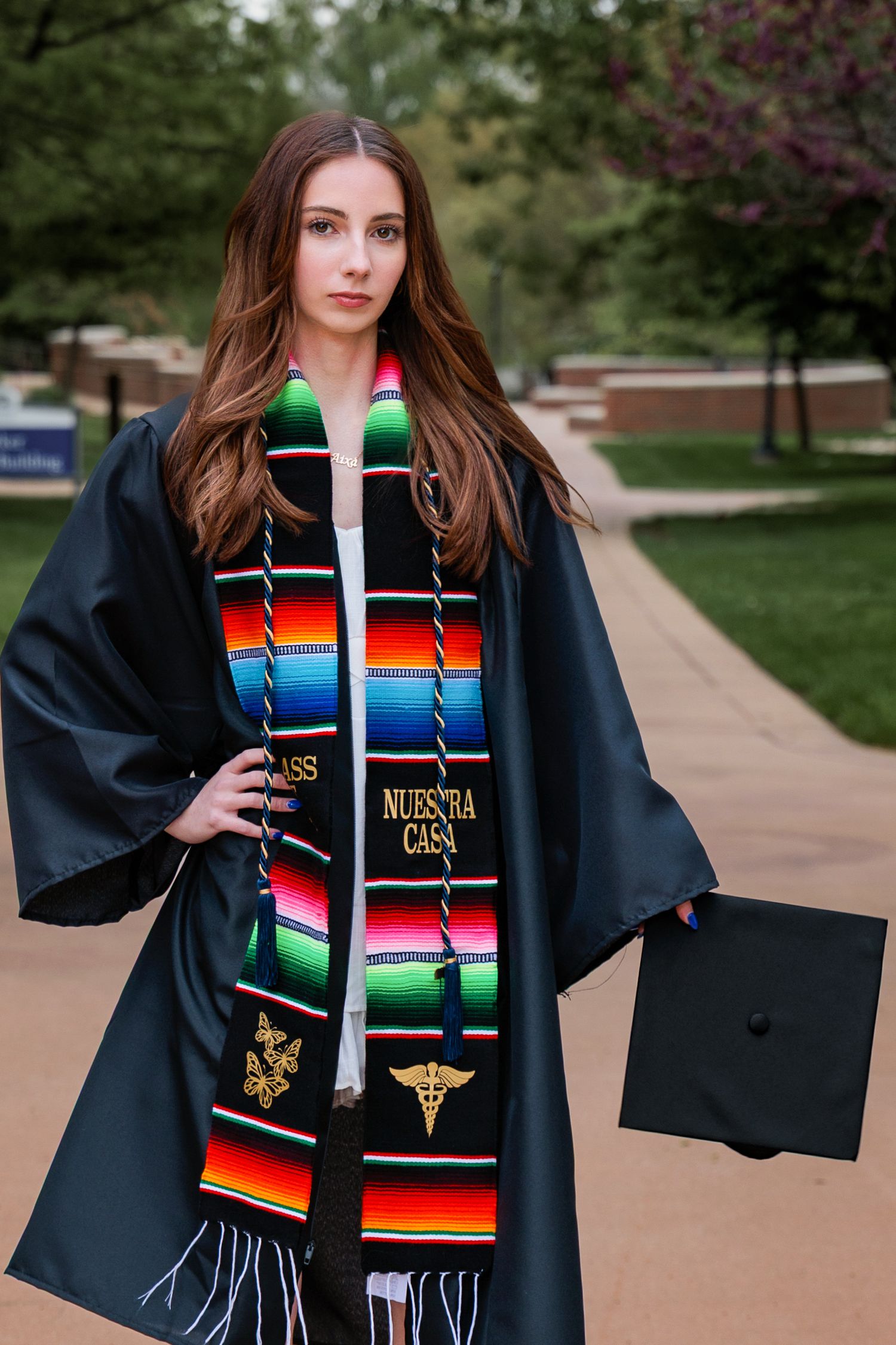 Graduate in black cap and gown with colorful Mexican serape-style graduation stole in outdoor campus setting.