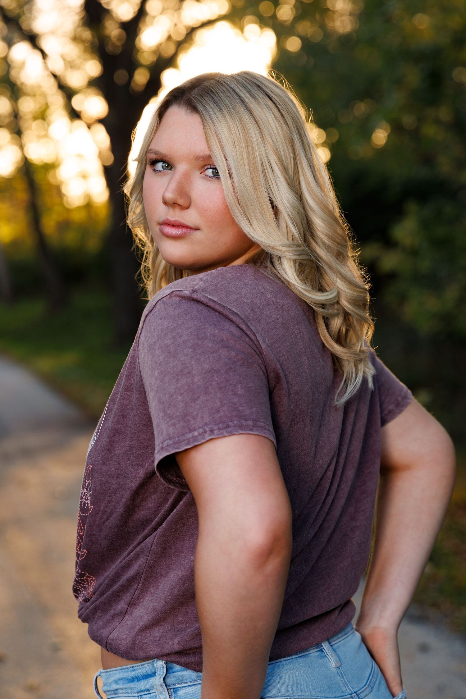A person wearing a brown t-shirt and jeans poses outdoors at sunset with golden bokeh lighting in the background.