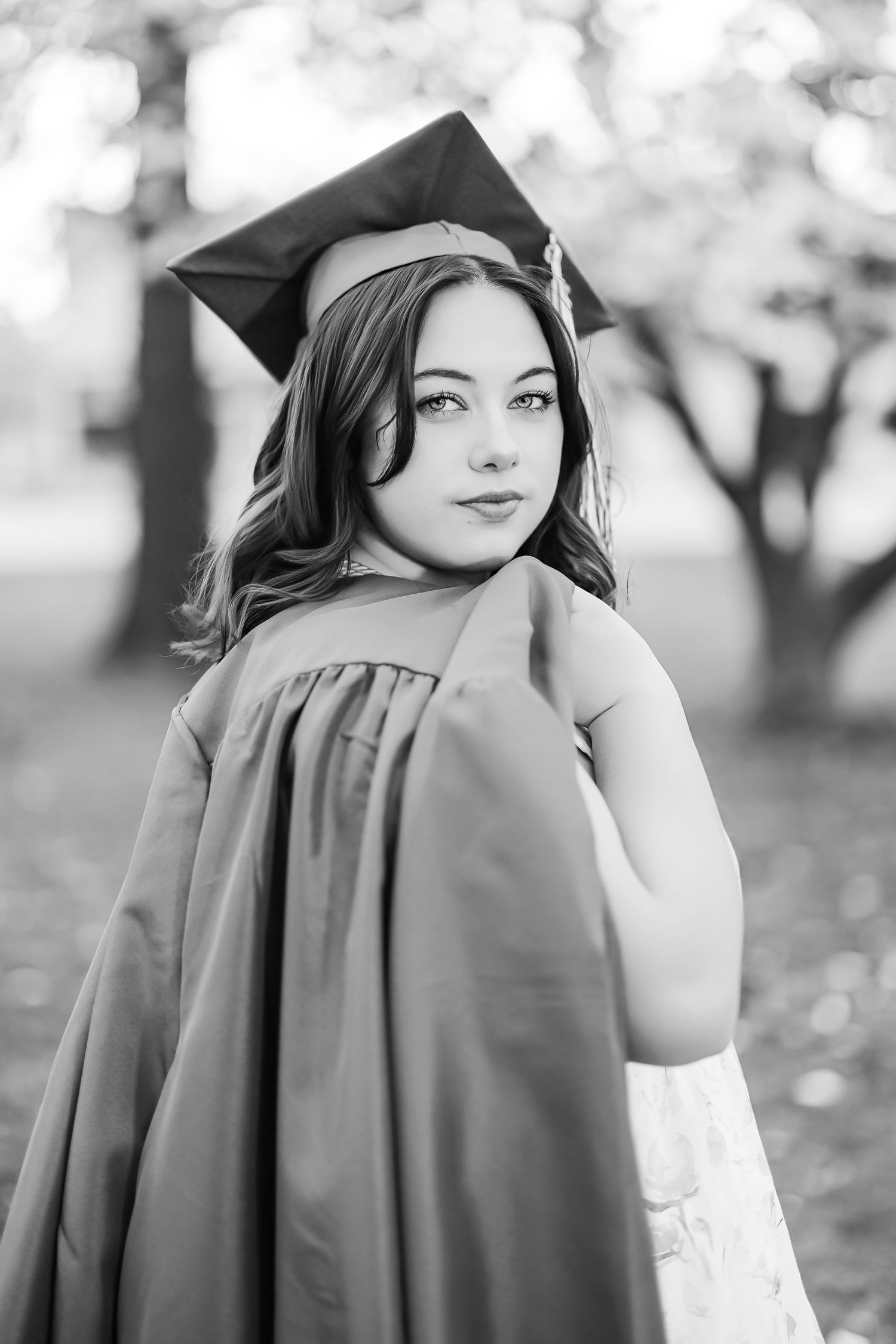 Black and white graduation portrait of a graduate in cap and gown looking over their shoulder outdoors.