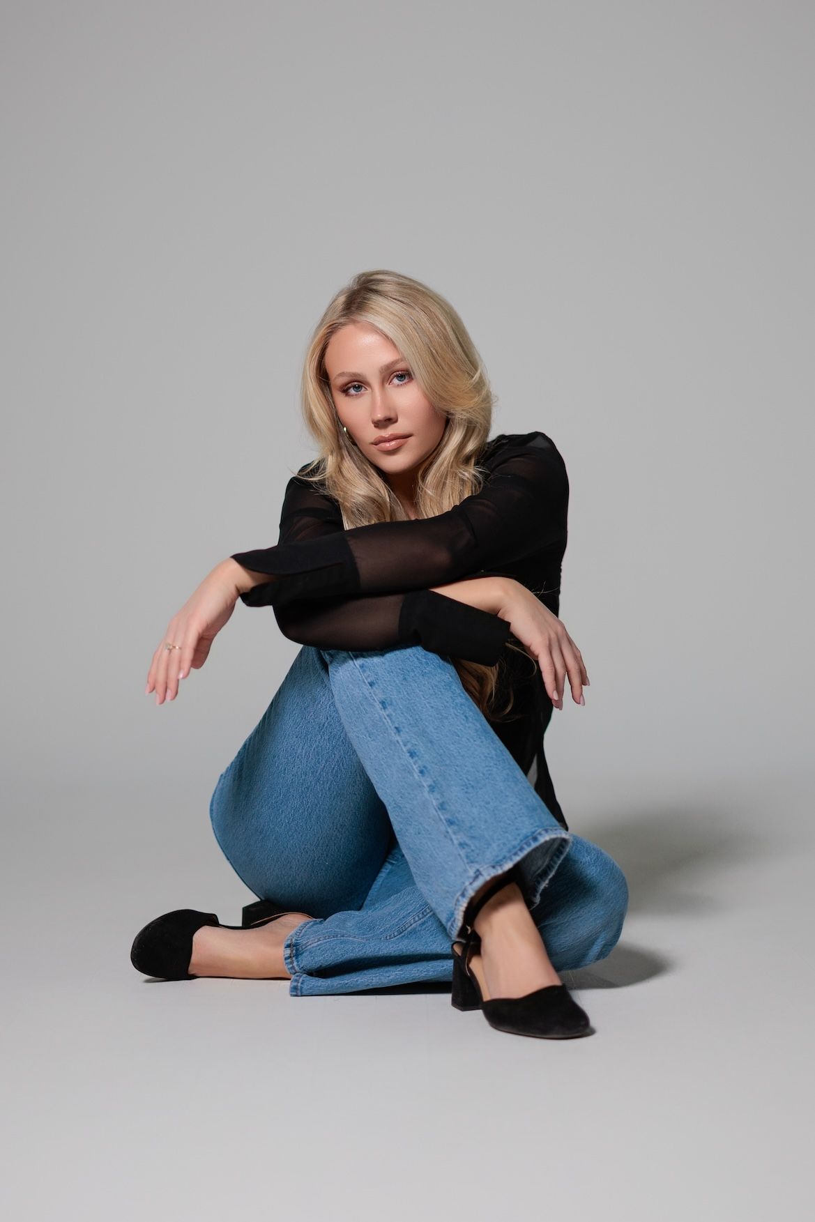 Casual fashion portrait with model in black top and blue jeans sitting on grey studio background.