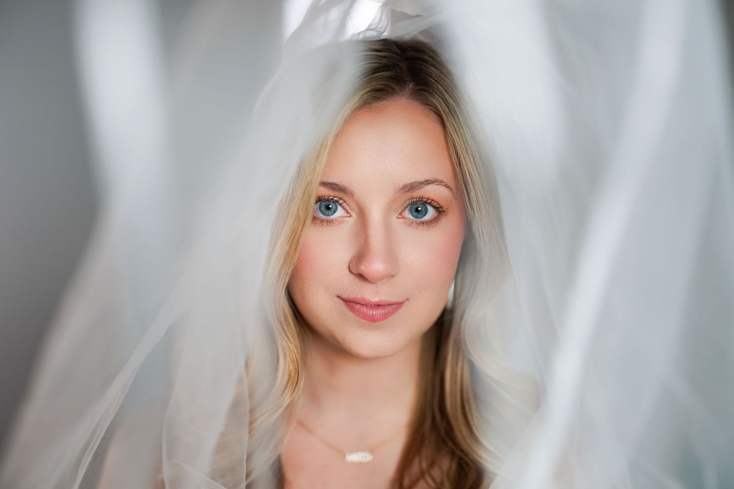 Bride with blonde hair wearing a white veil gazes softly at camera with natural makeup and soft lighting.