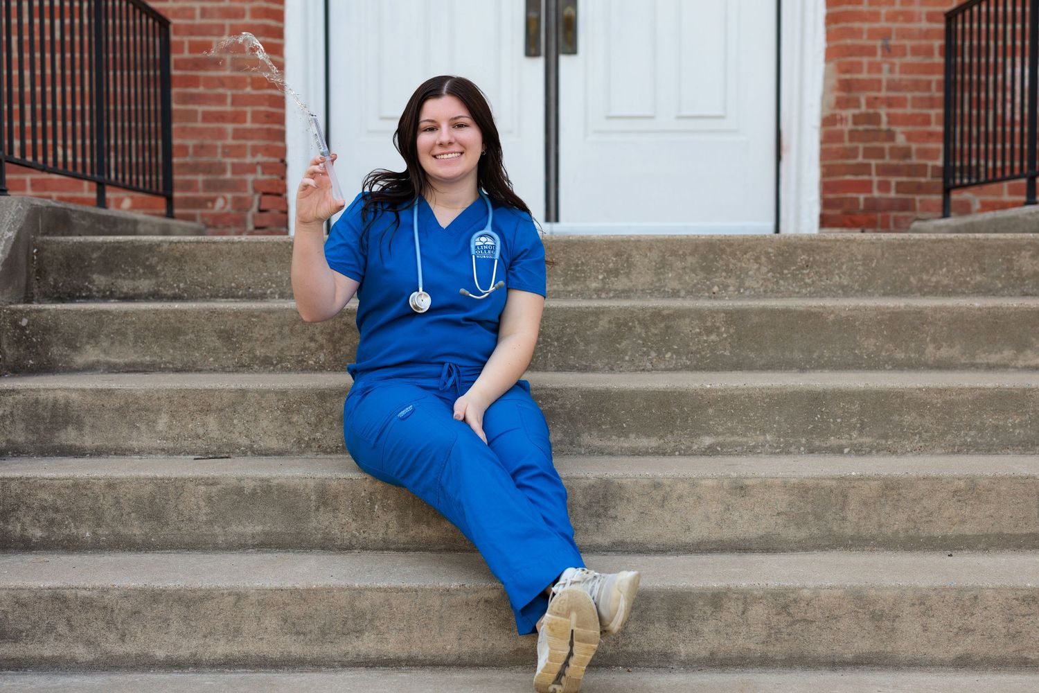 Medical professional in blue scrubs sits on concrete steps while giving a thumbs up outside a brick building.