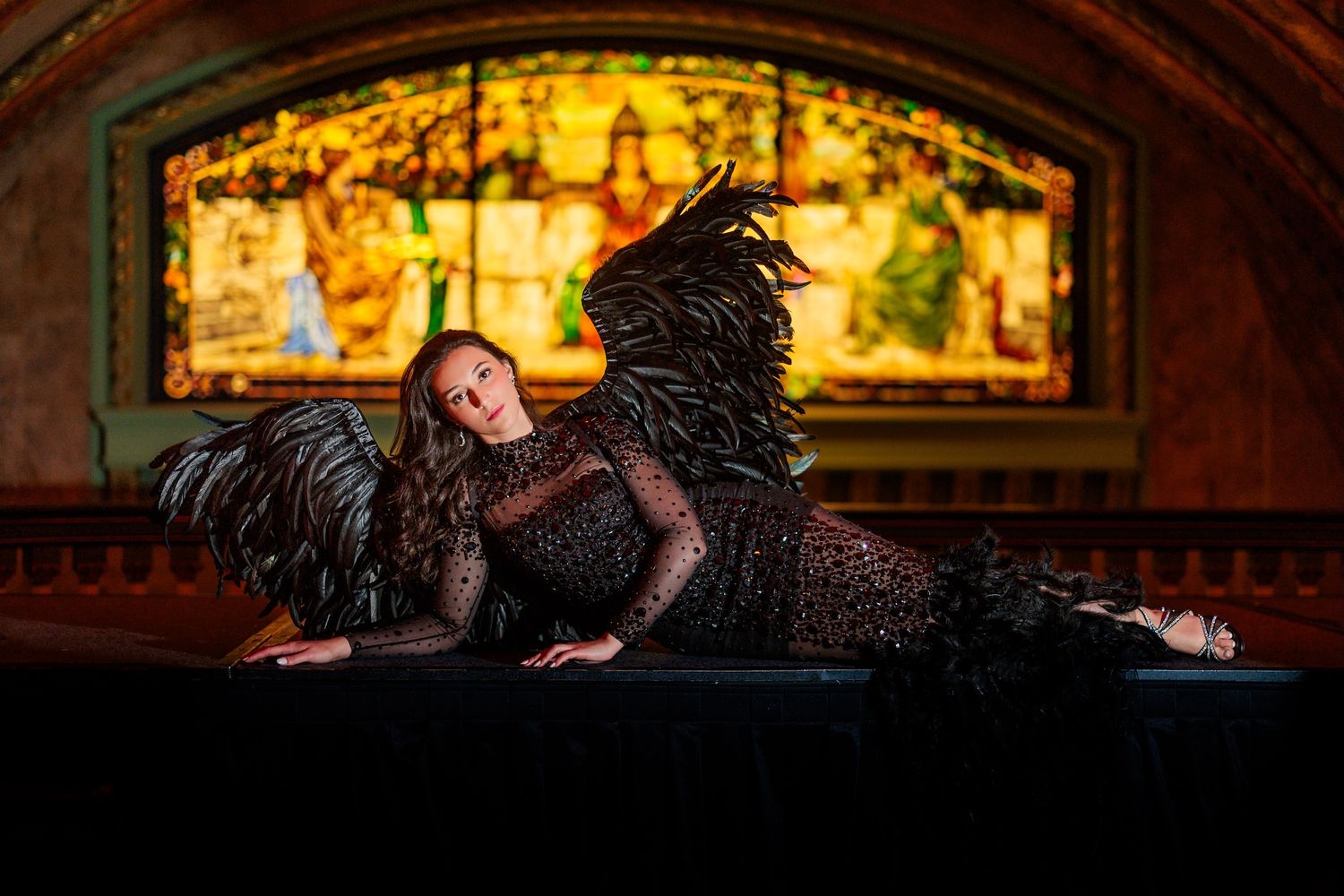 A dramatic photo of a dancer in black feathers reclining on stage with stained glass windows glowing in the background.