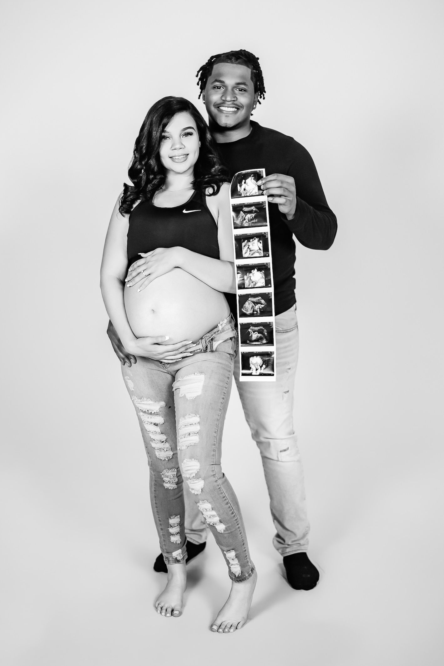 Black and white maternity photo showing expectant couple displaying ultrasound strip while standing against studio backdrop.