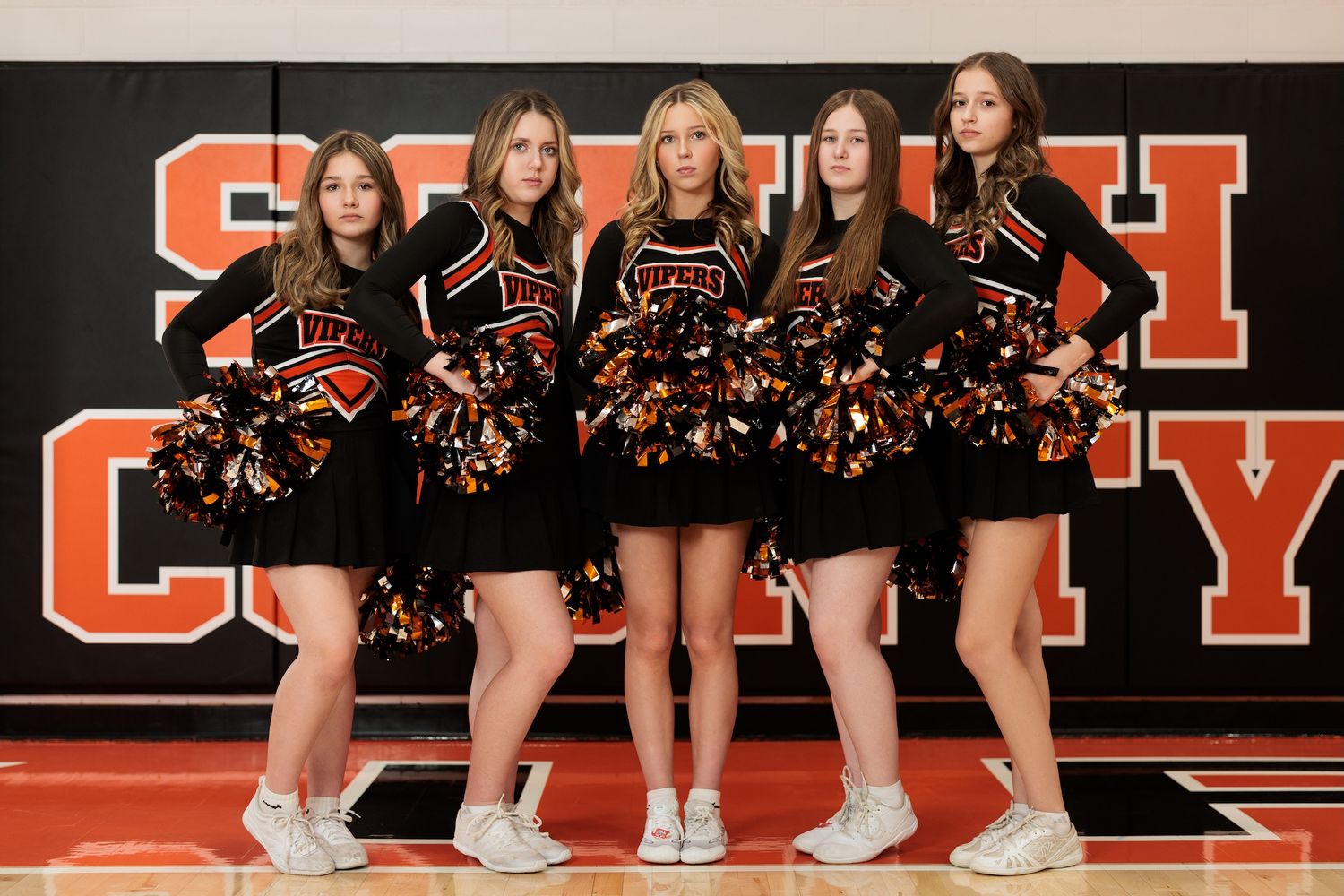 Five cheerleaders in black and orange uniforms pose together on a basketball court with school branding.
