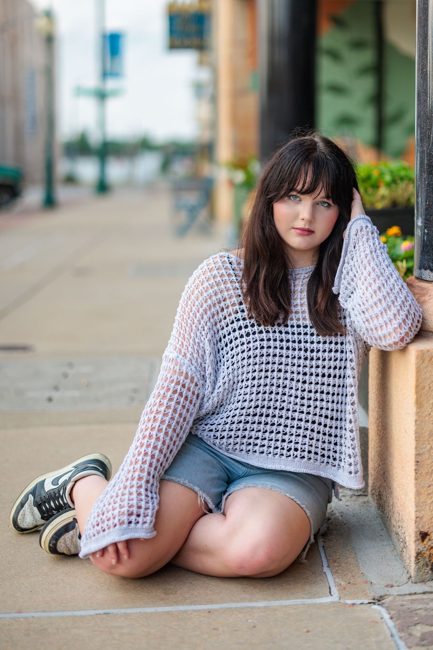 A person in a white crochet sweater and denim shorts sits on a sidewalk wearing sneakers.