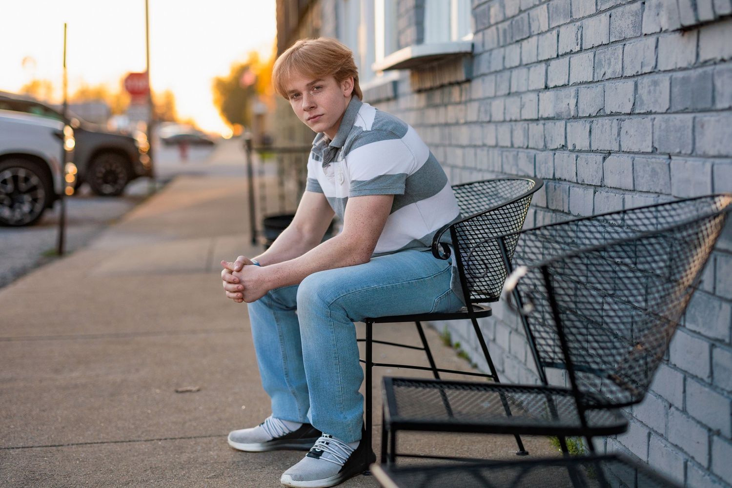 Someone wearing a striped polo shirt and jeans sits thoughtfully on a bench against a brick wall at sunset.