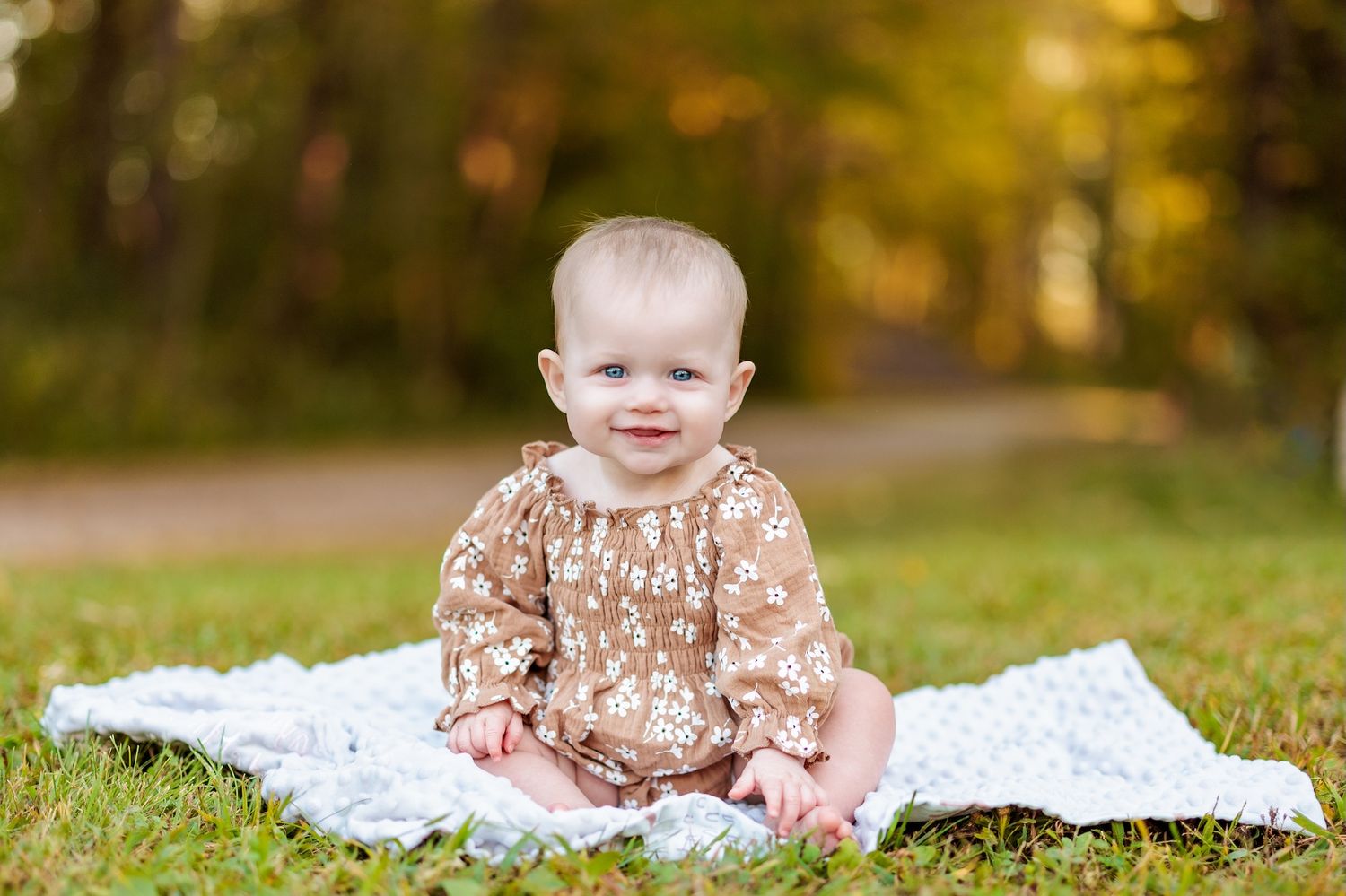 Baby in a floral brown dress sits smiling on a white blanket outdoors during autumn.