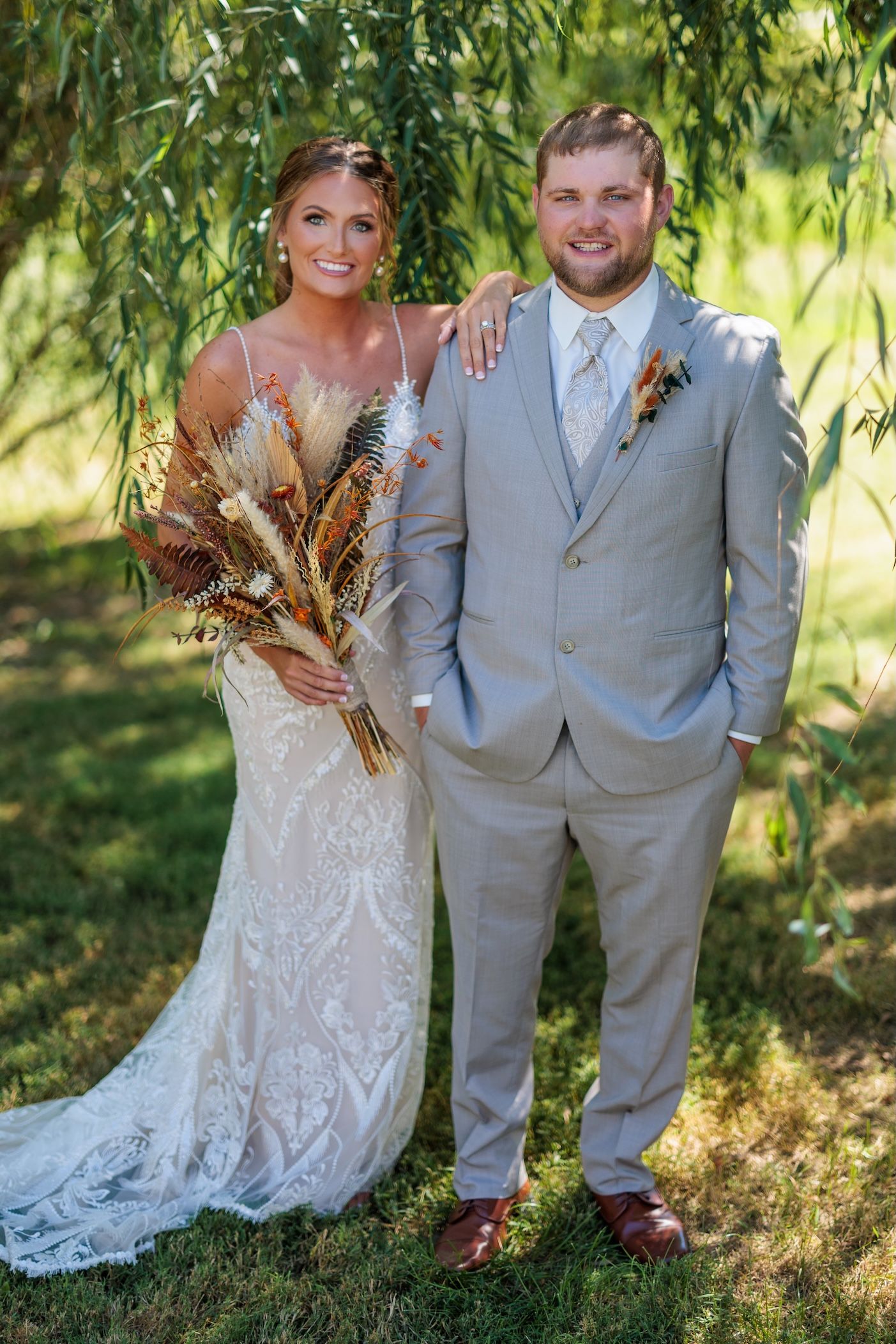 A wedding couple poses beneath a willow tree, with the bride holding a dried pampas grass bouquet.