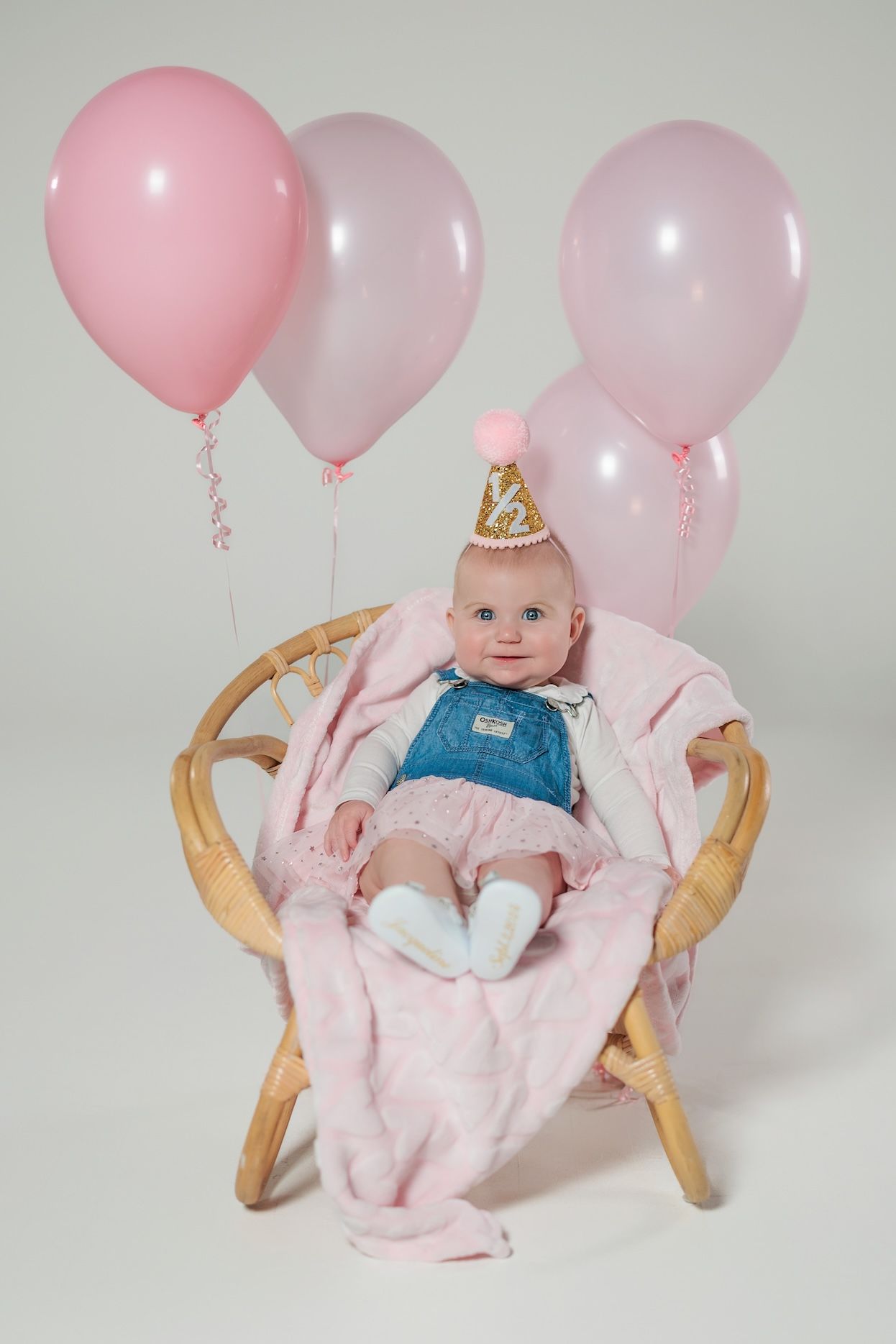 Baby in party hat seated in rattan chair with pink and white balloons for first birthday celebration.