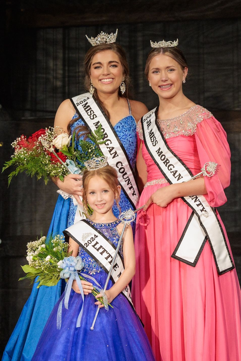 Three pageant winners in elegant gowns and crowns pose together while holding bouquets at an awards ceremony.