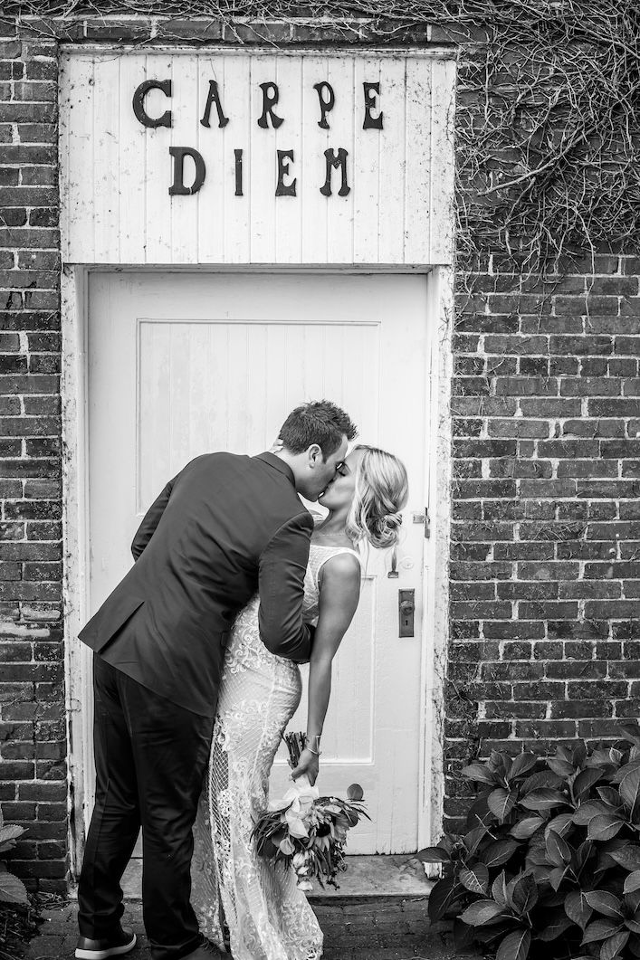 A couple shares a romantic kiss in front of a white door with 'Carpe Diem' text above it on a brick wall.
