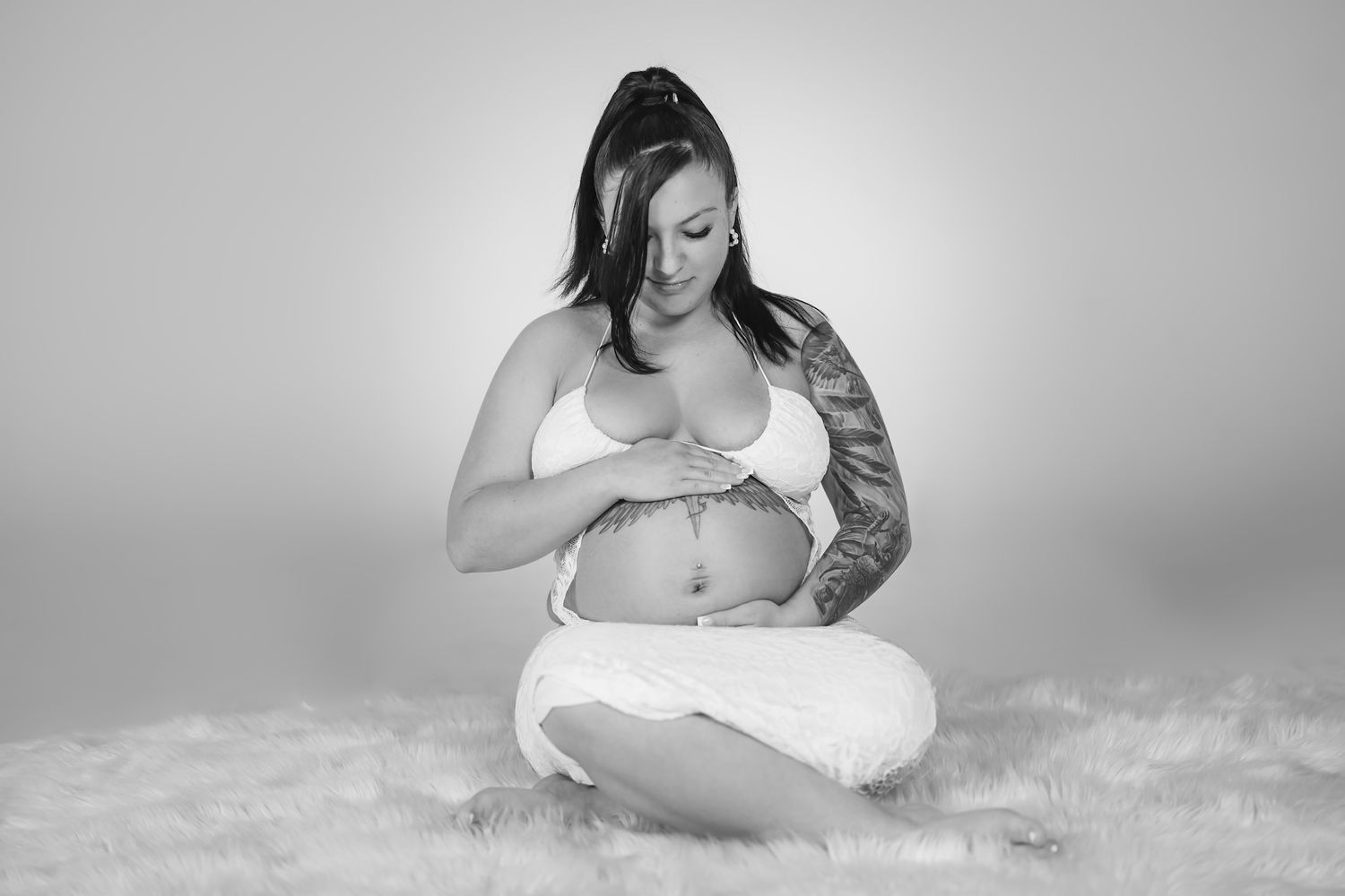 Black and white maternity portrait of an expectant mother sitting on a plush rug in intimate white attire.