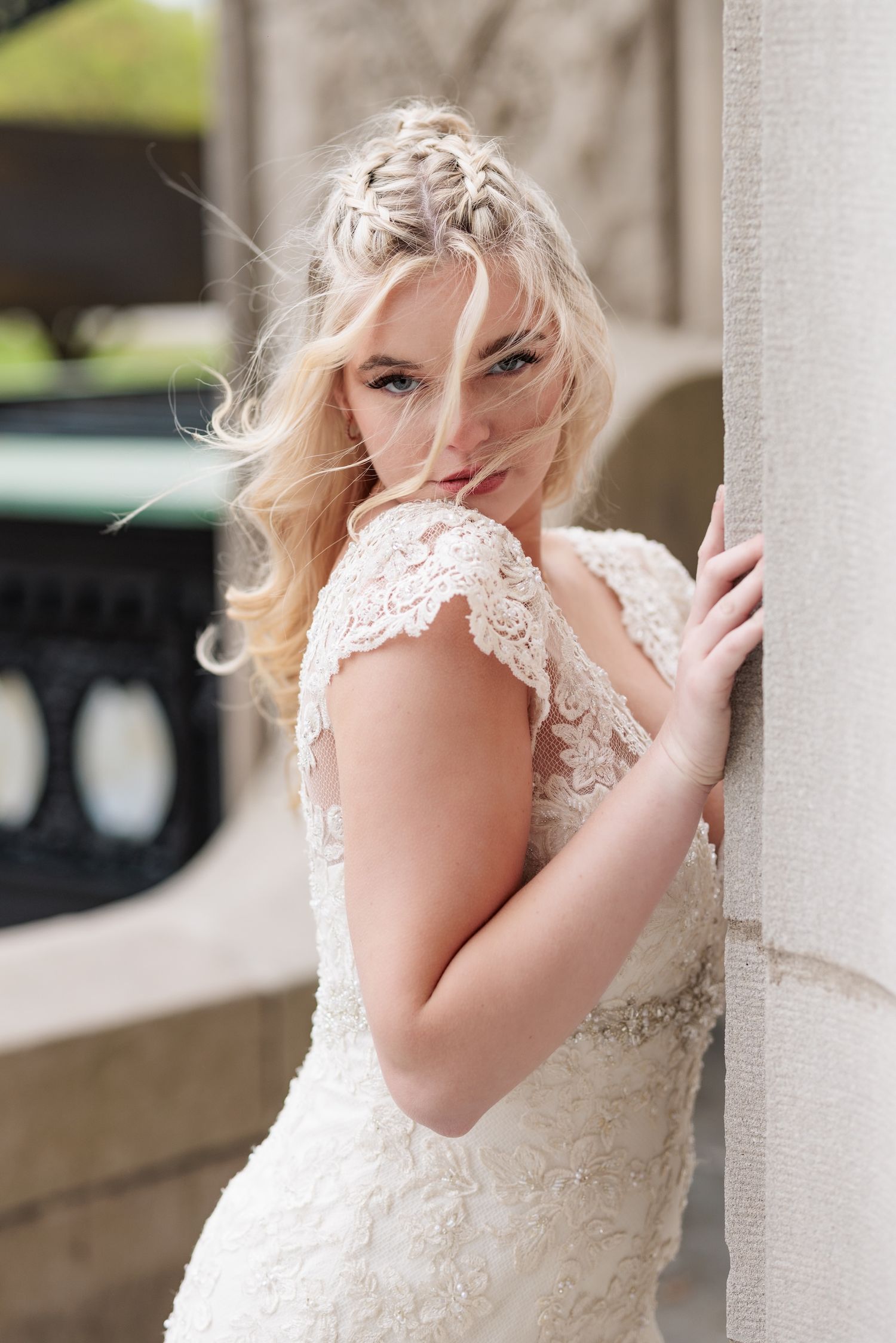 Elegant bride in lace wedding dress with cap sleeves poses playfully against white pillar outdoors.
