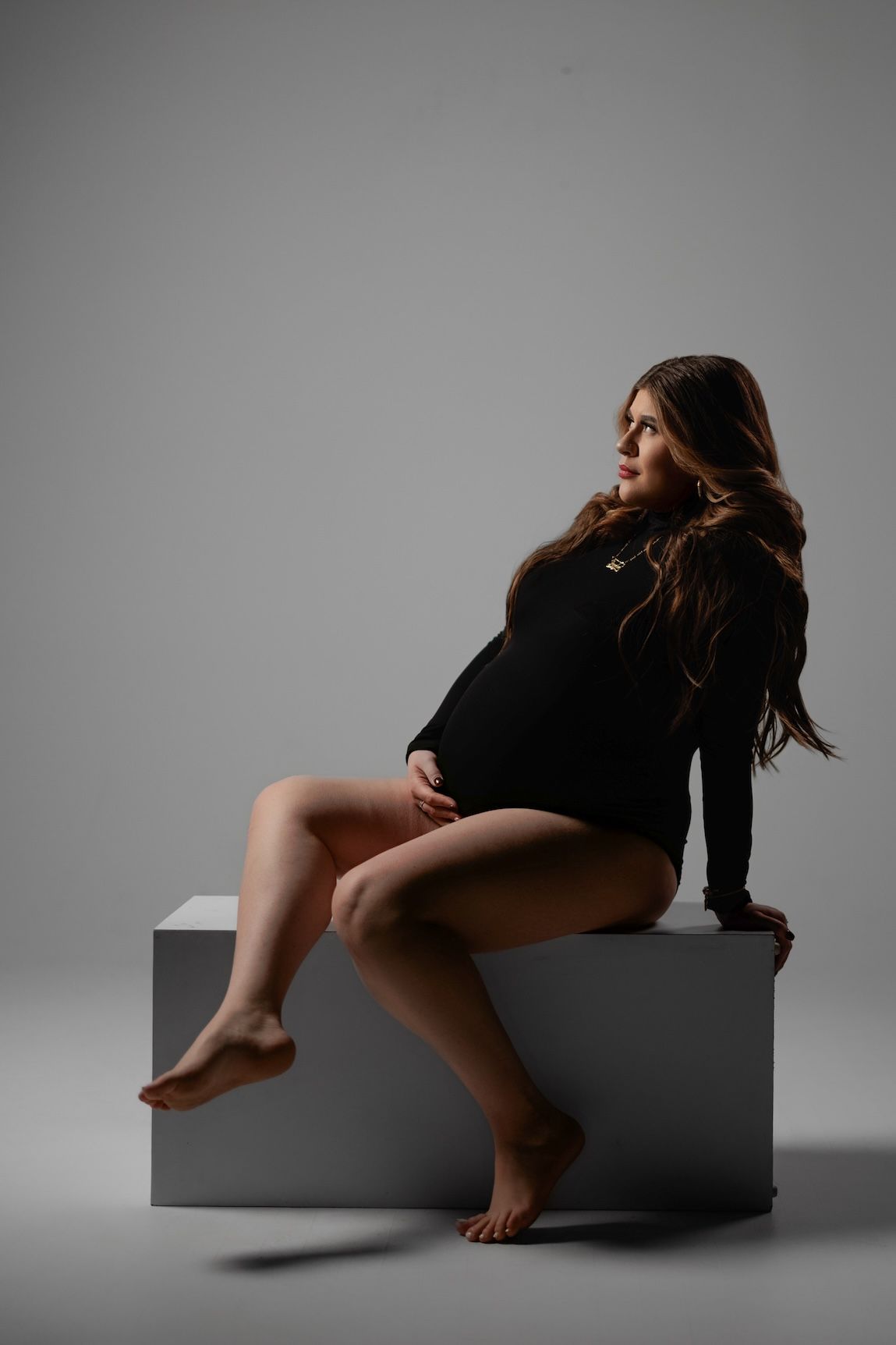 Artistic maternity photo with silhouette seated on cube against gray backdrop in black bodysuit.