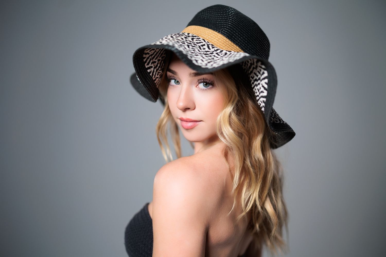 Portrait shot of someone wearing a patterned black and white hat with blonde wavy hair against a gray background.