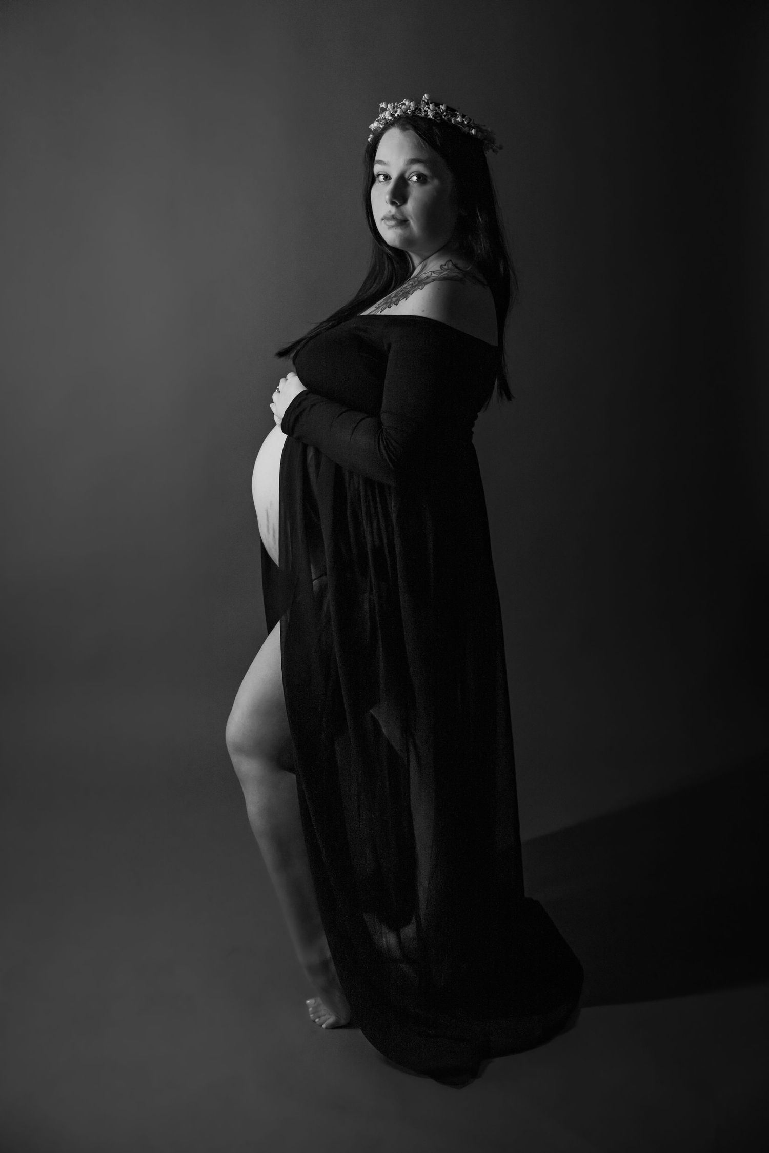 A dramatic black and white maternity photo featuring an off-shoulder long black dress and floral crown in silhouette.