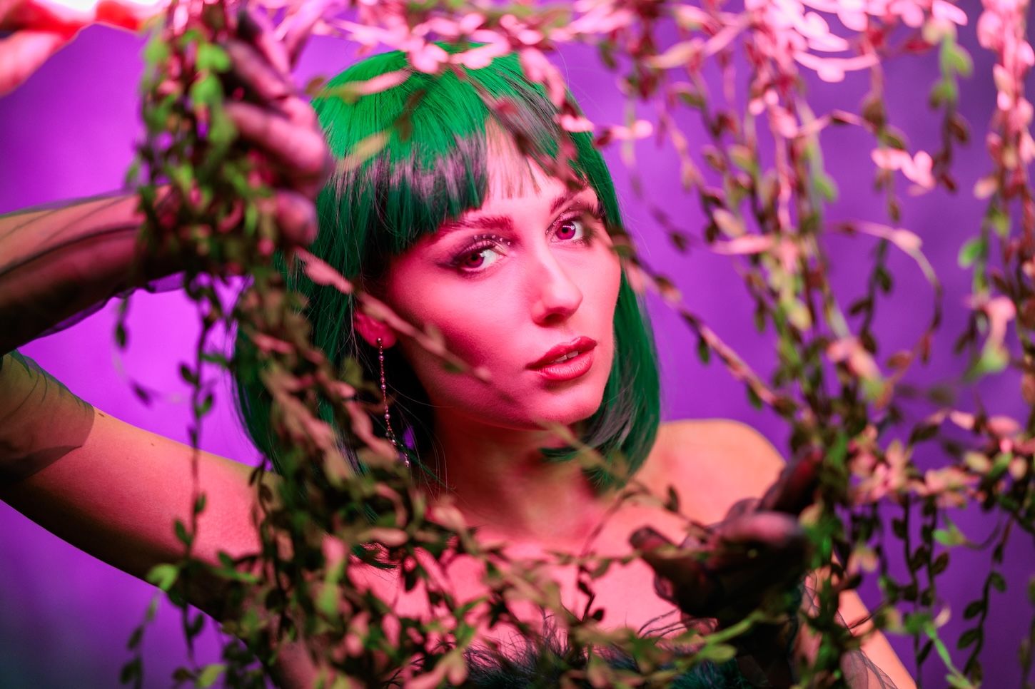 Artistic portrait with green hair and pink lighting through floral branches, creating a dreamy atmosphere.