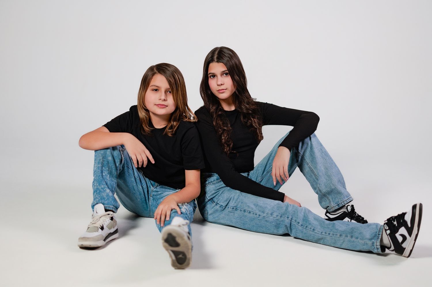 Two individuals in matching black t-shirts and blue jeans strike casual poses while sitting on studio floor.