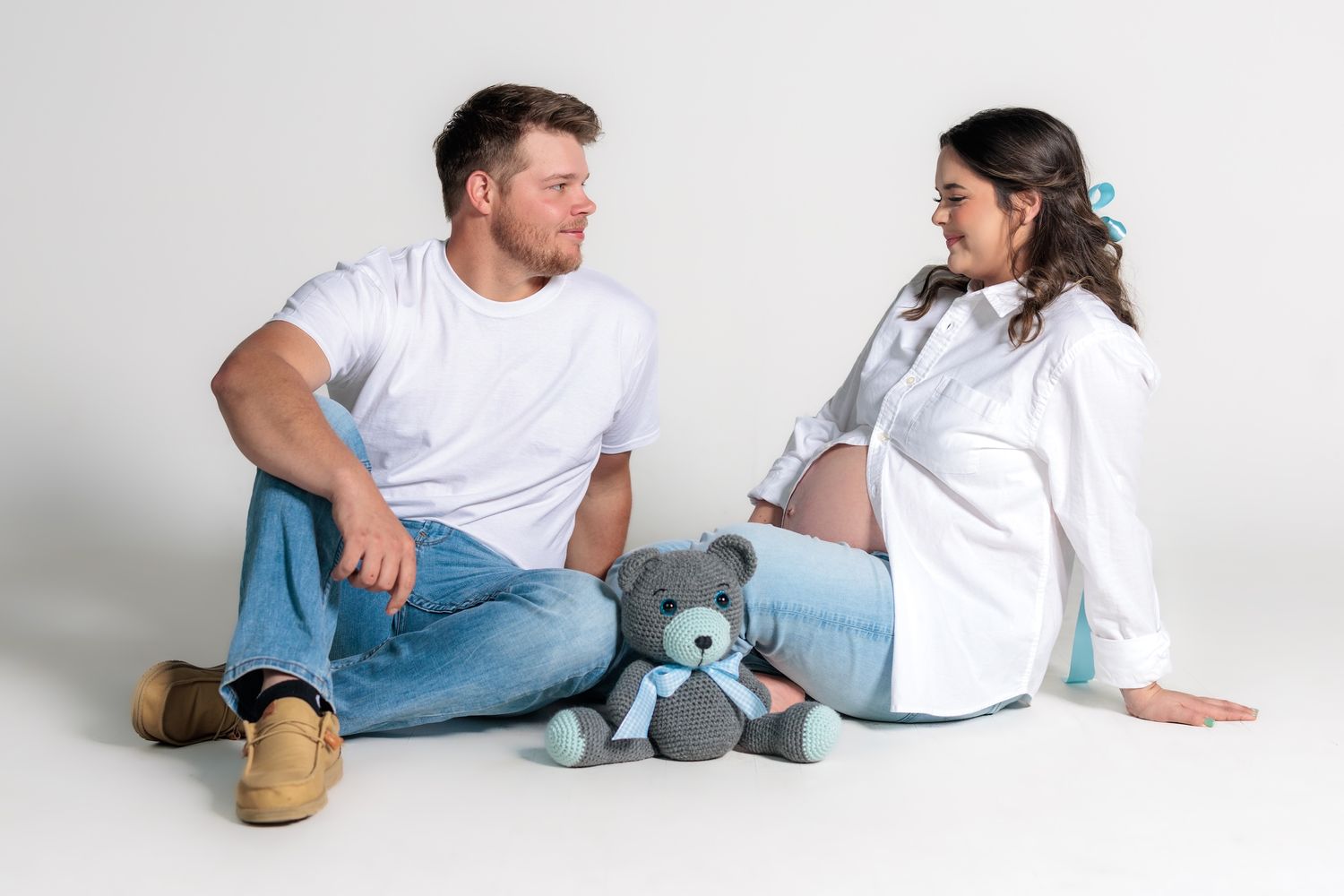 An expectant couple sits casually on the floor with a teddy bear in a minimalist studio setting.