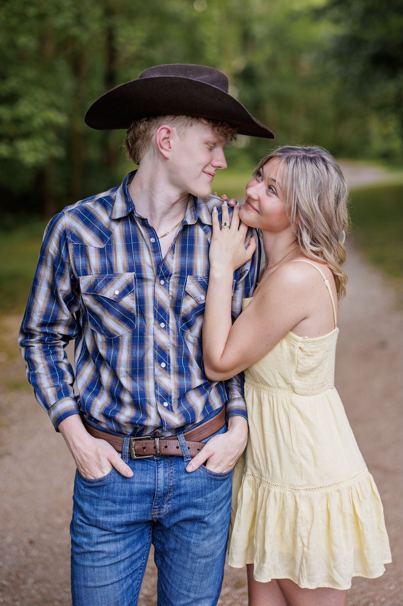 A couple poses on a path outdoors, with him in western wear and her in a yellow sundress.