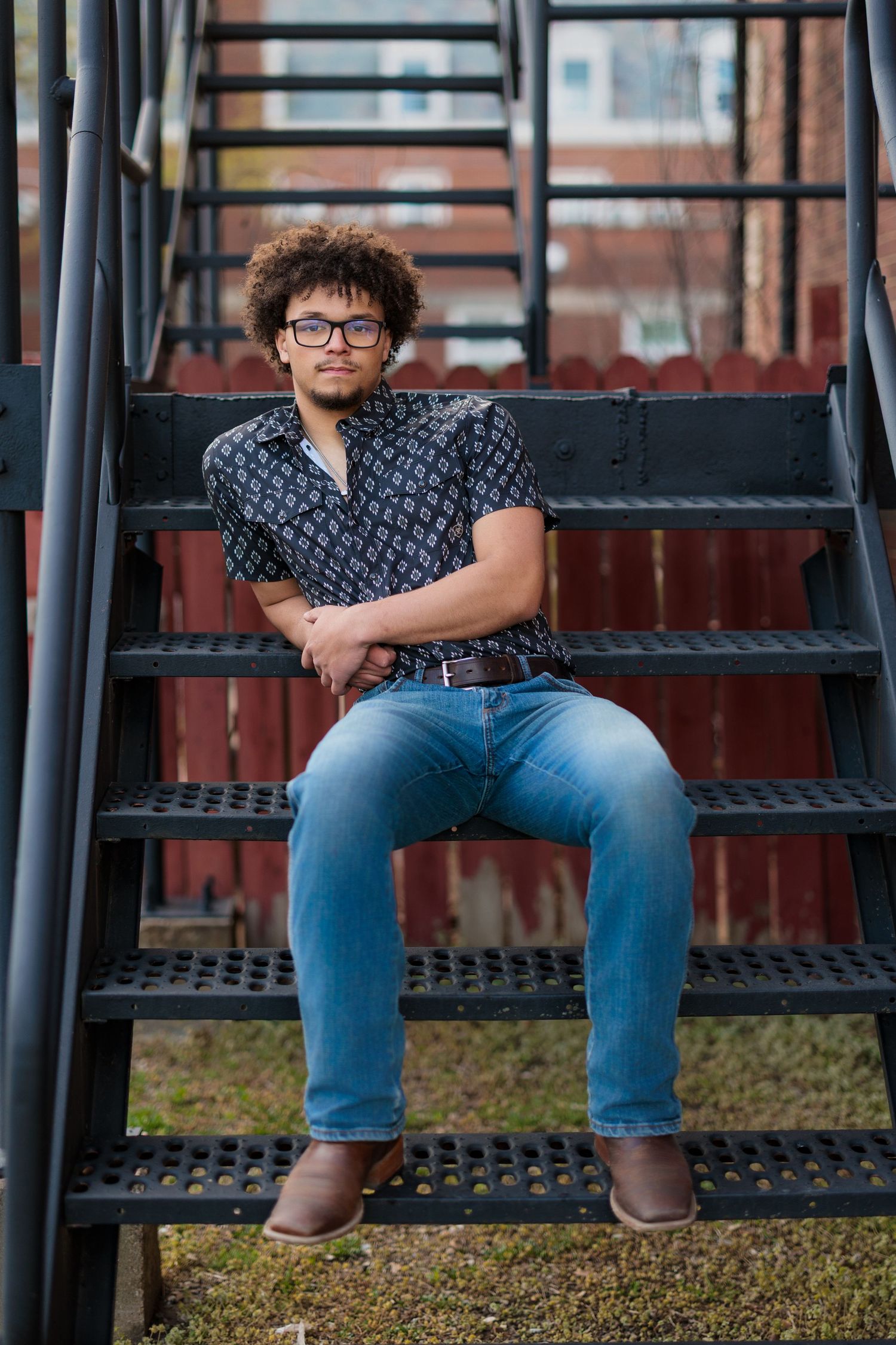 A person in glasses and patterned shirt sits on metal outdoor stairs wearing blue jeans and brown shoes.