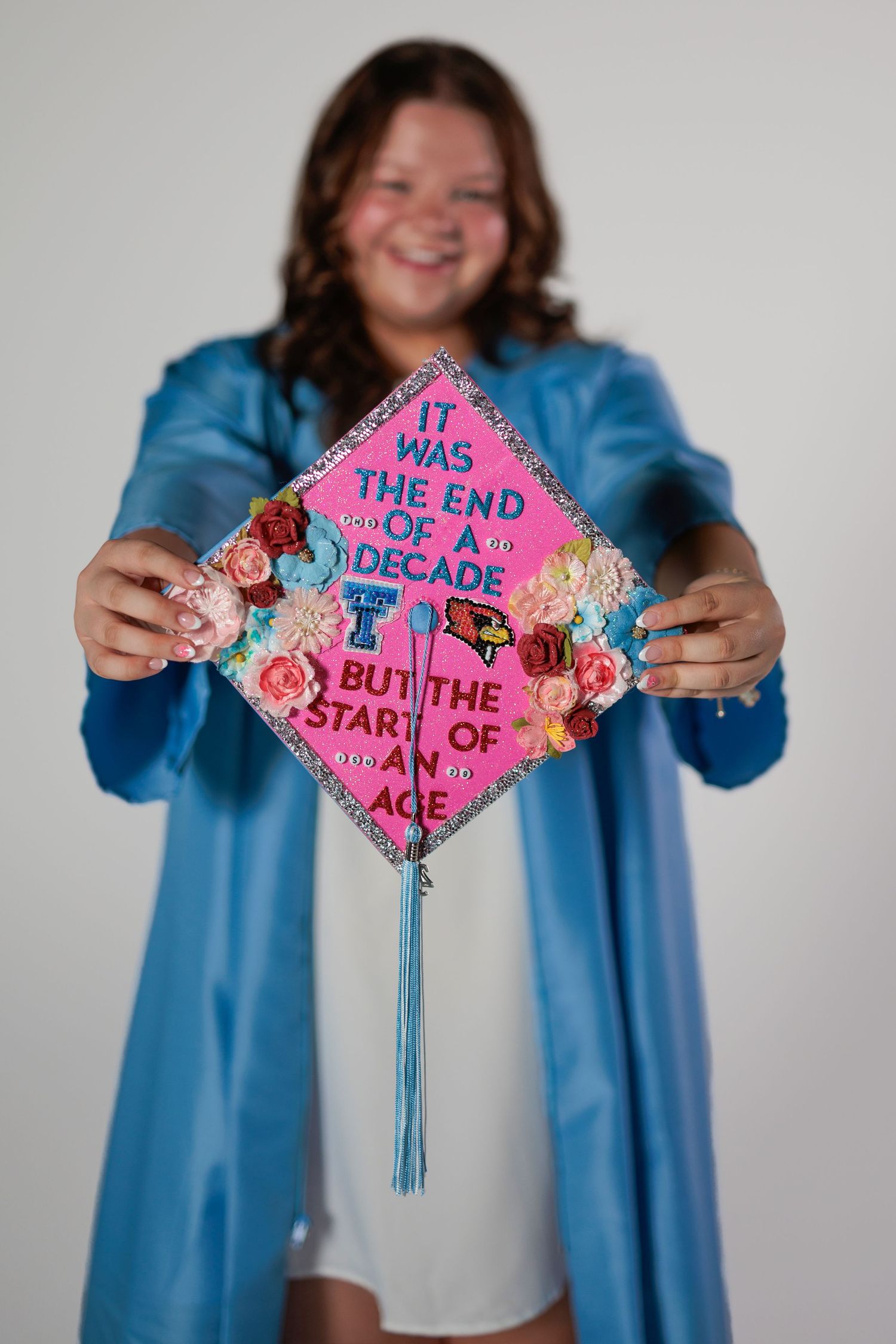 A decorated pink graduation cap with floral designs and tassels is held up against a blue gown backdrop.