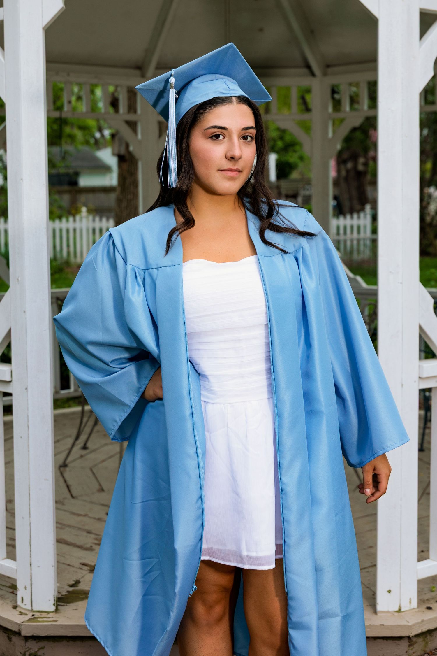 Graduate wearing light blue cap and gown stands confidently in white gazebo during commencement ceremony.