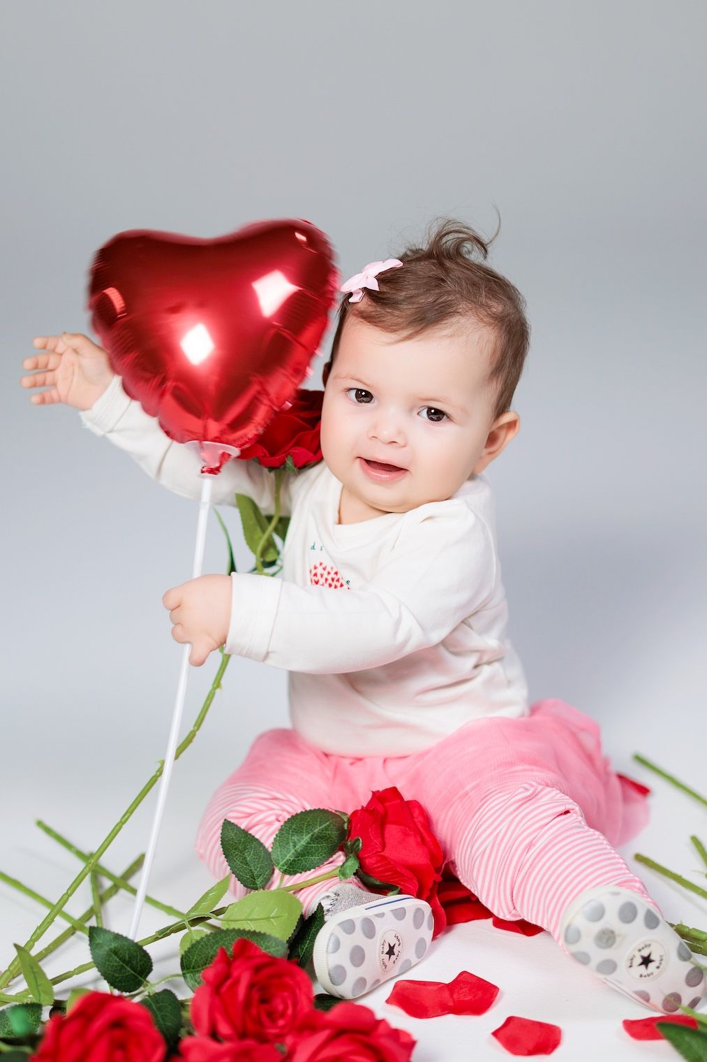 Child in white and pink outfit sitting with red roses and heart balloon for Valentine's Day photo.