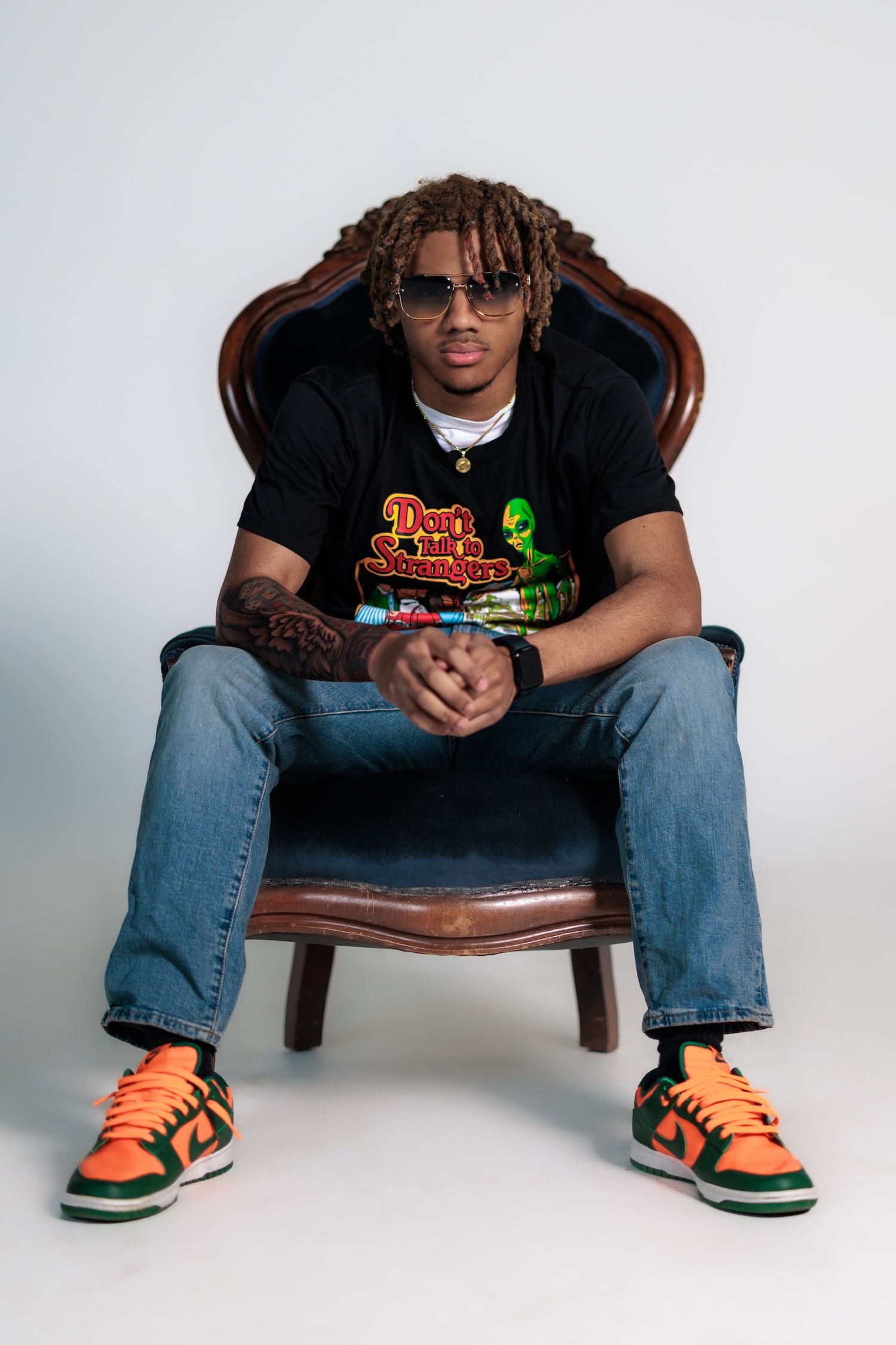 Person in orange and green Nike sneakers sits in vintage leather chair wearing black t-shirt and denim jeans.