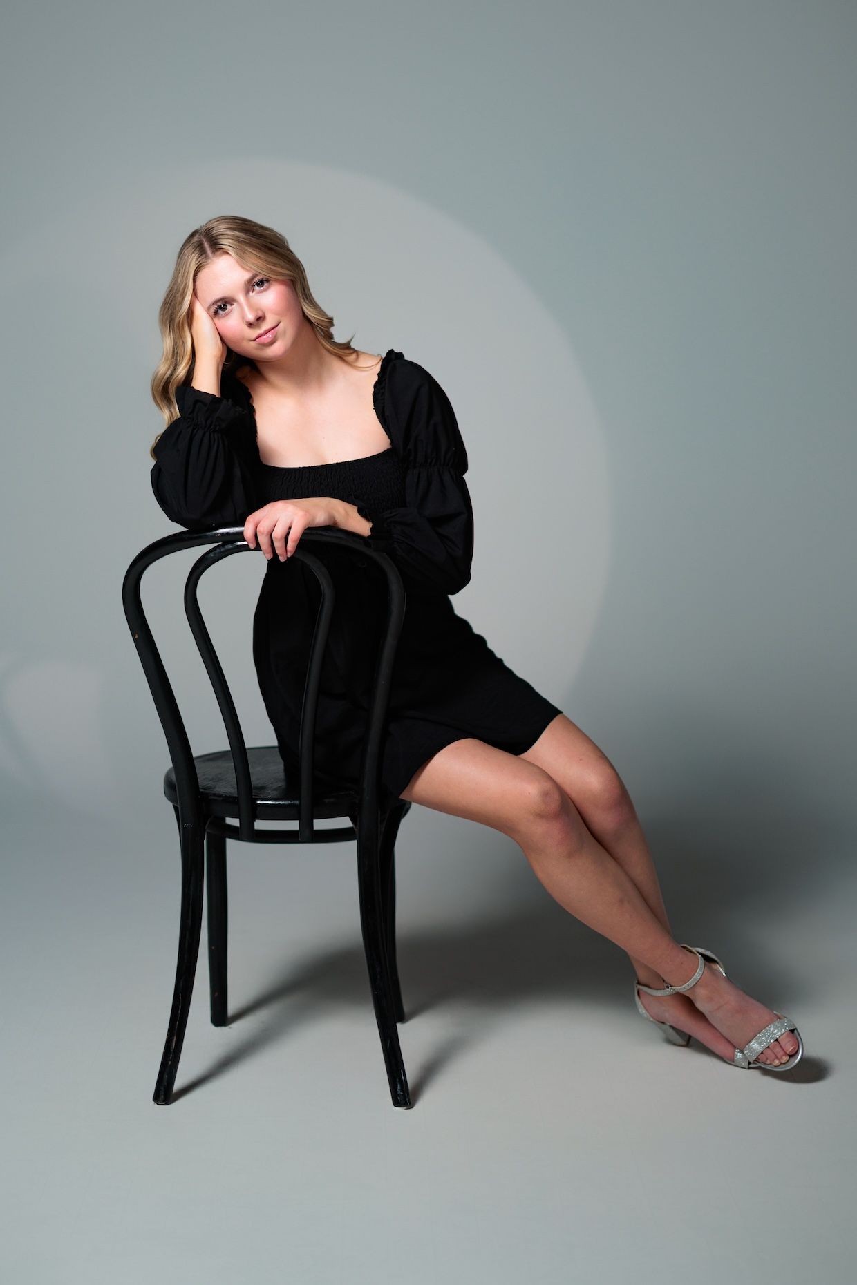Studio portrait with person in black dress seated on classic bentwood chair against gray background, wearing silver sandals.