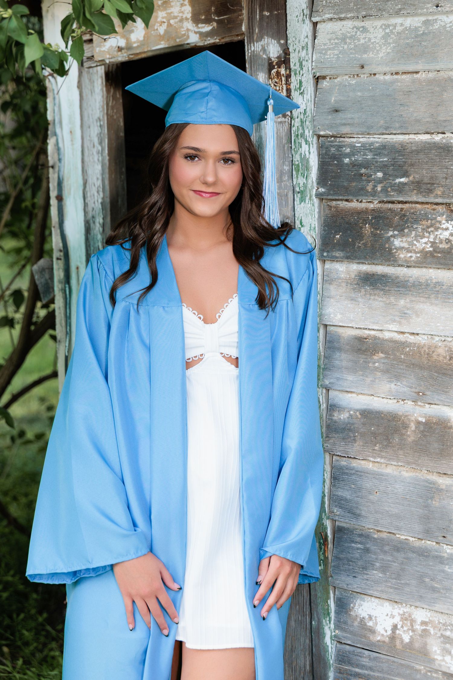 Graduate wearing light blue cap and gown stands against rustic wooden wall with white dress underneath.