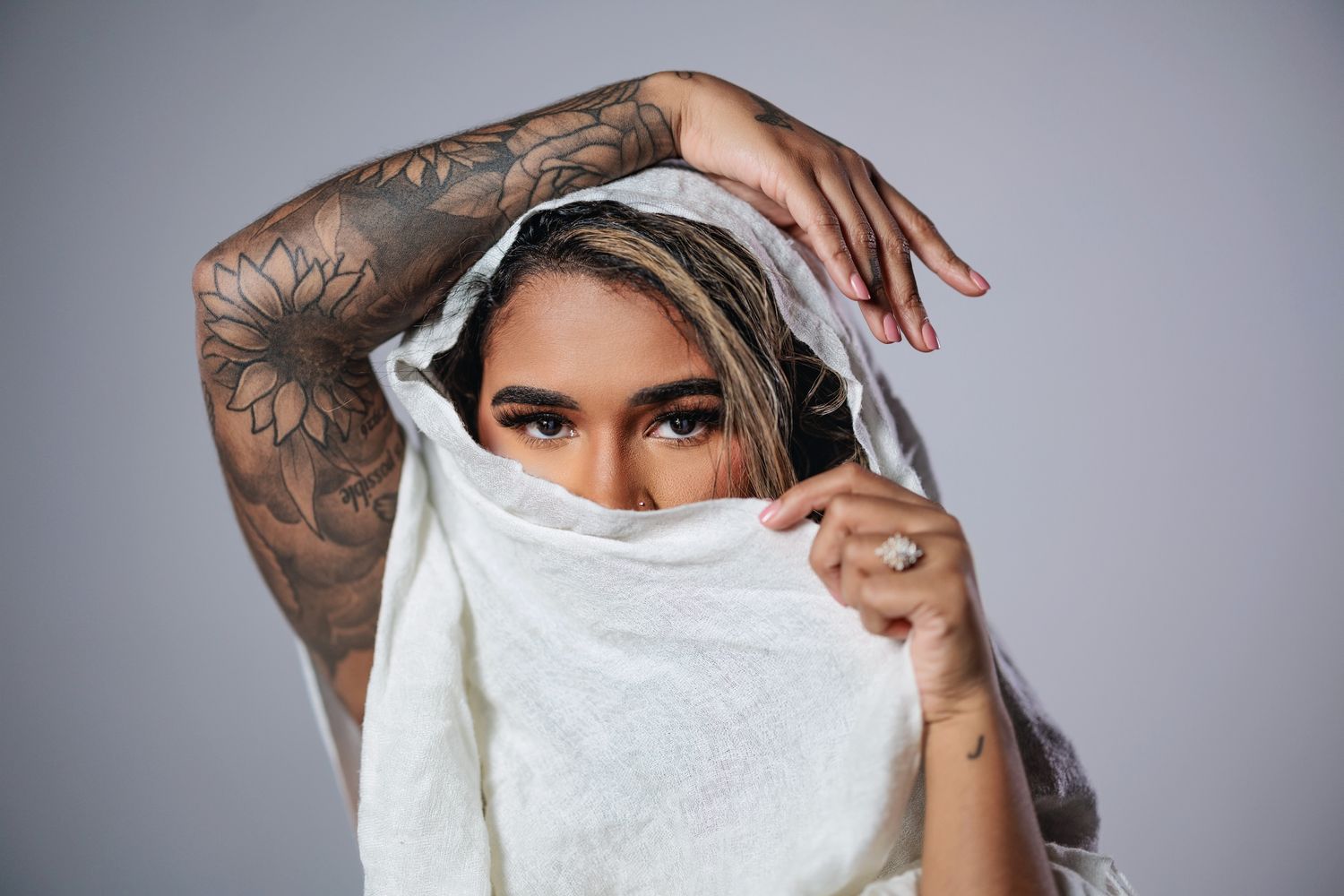 A person with floral tattoos on their arm poses with a white cloth partially covering their face.