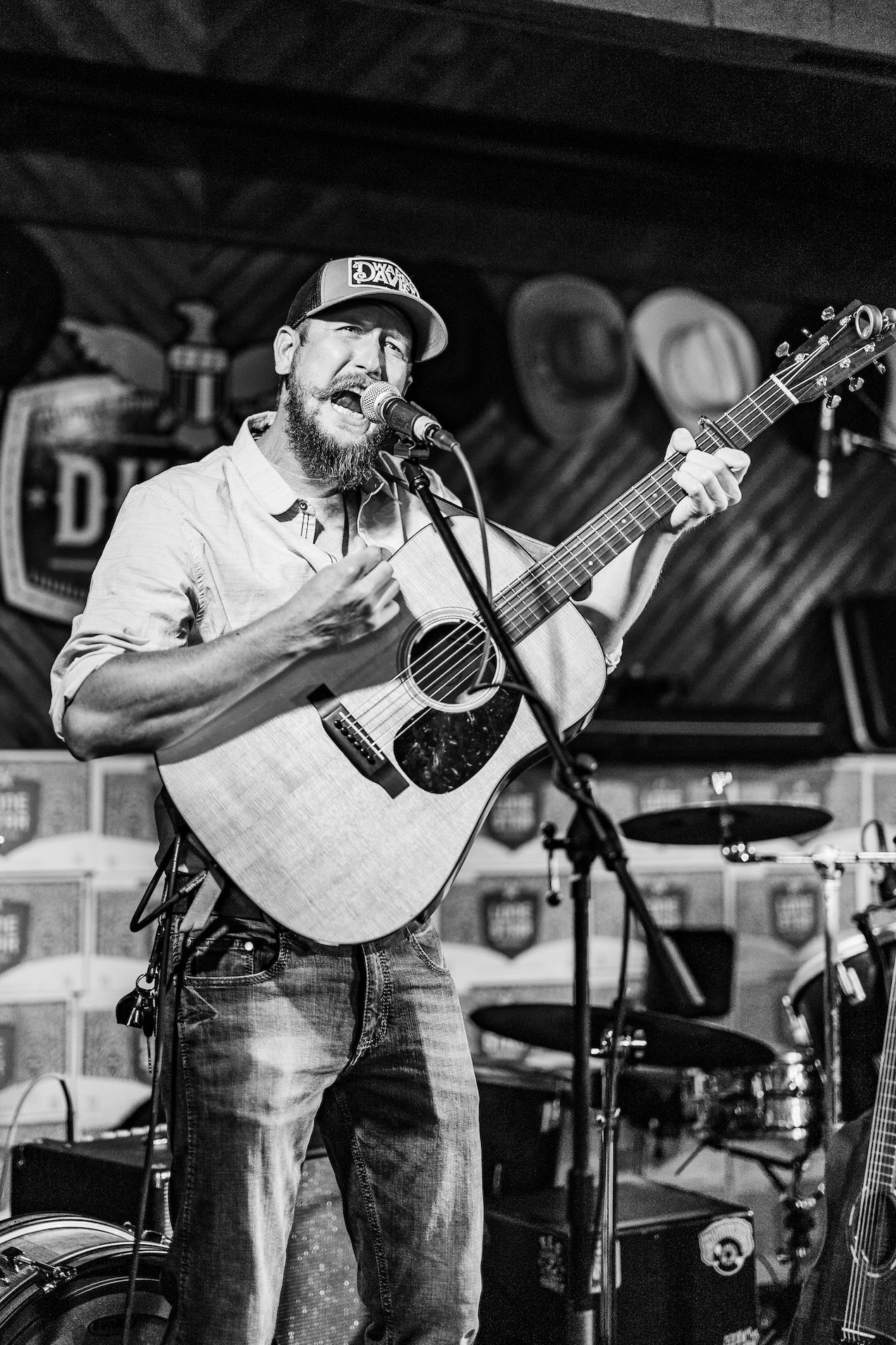 Black and white concert photo of a musician in baseball cap playing acoustic guitar and singing into microphone.