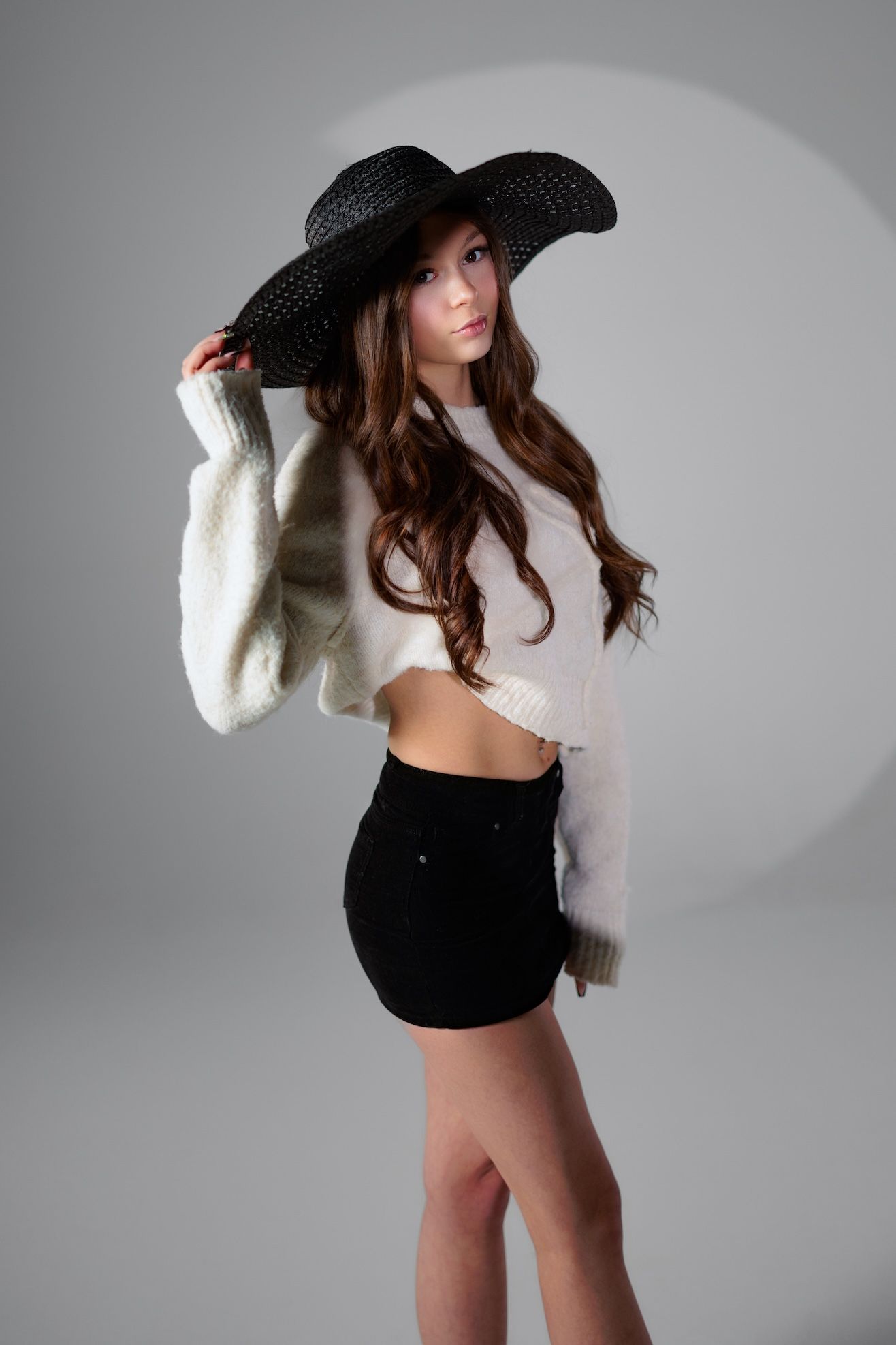 Fashion photo of model in black wide-brimmed hat, white cropped sweater and black shorts against grey background.